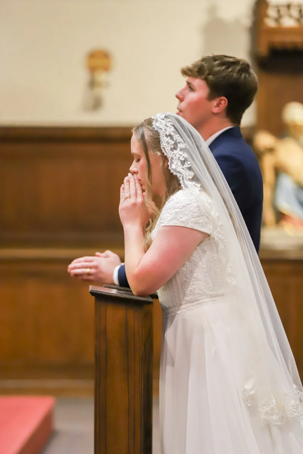 A bride and groom praying during their wedding ceremony in a church, with hands clasped and eyes closed.