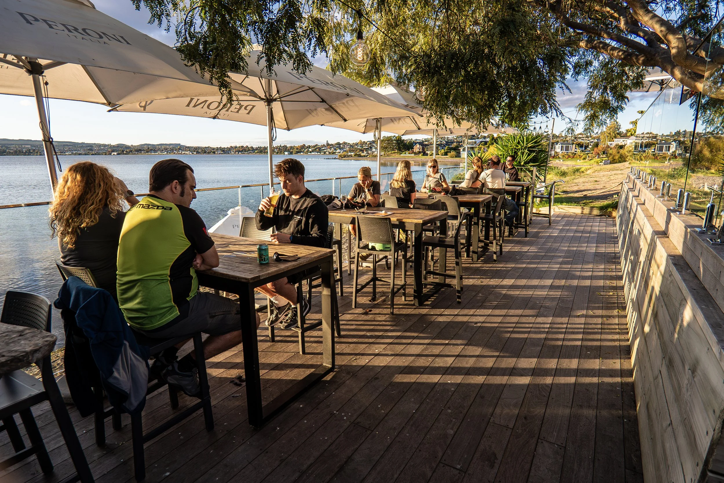 People sitting at outdoor tables on a deck by the water, under umbrellas, with trees and a cityscape in the background, enjoying drinks during the daytime.