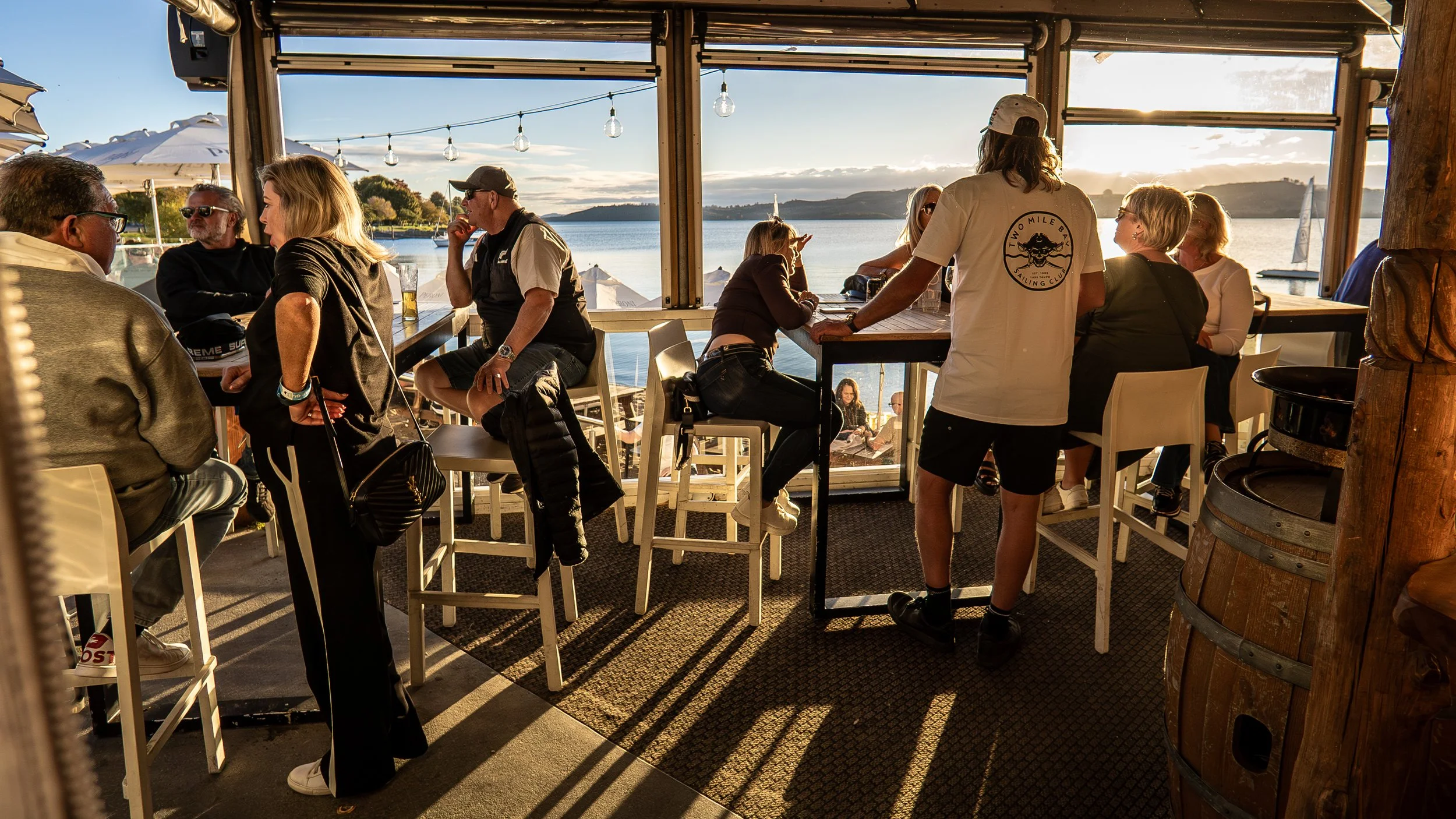 People sitting and standing at a waterfront bar during sunset, enjoying drinks and conversation with sailboats in the background.