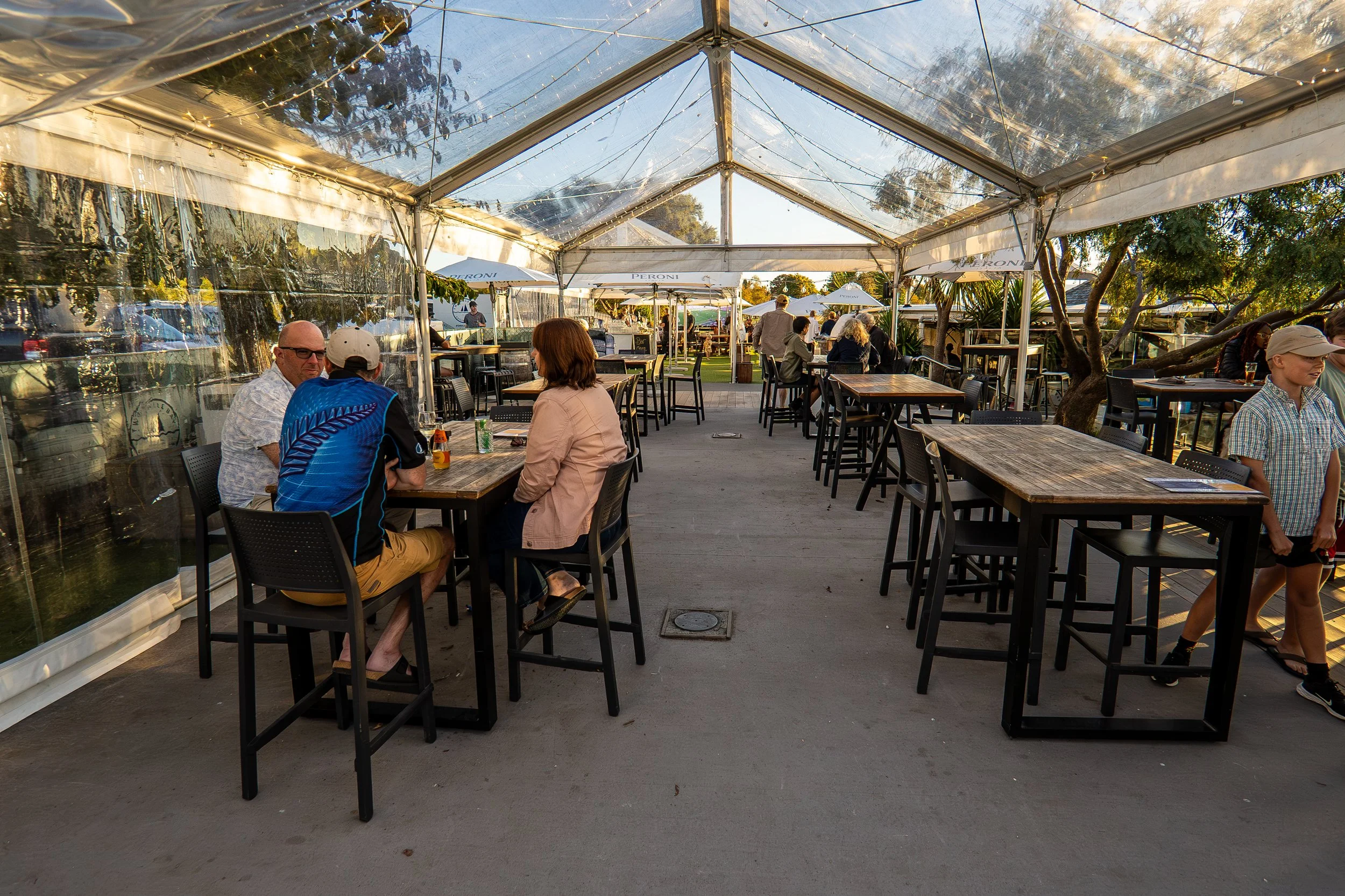 Outdoor dining area under a clear tent with wooden tables and black chairs, some people sitting and chatting, trees and umbrellas in the background, warm late afternoon lighting.