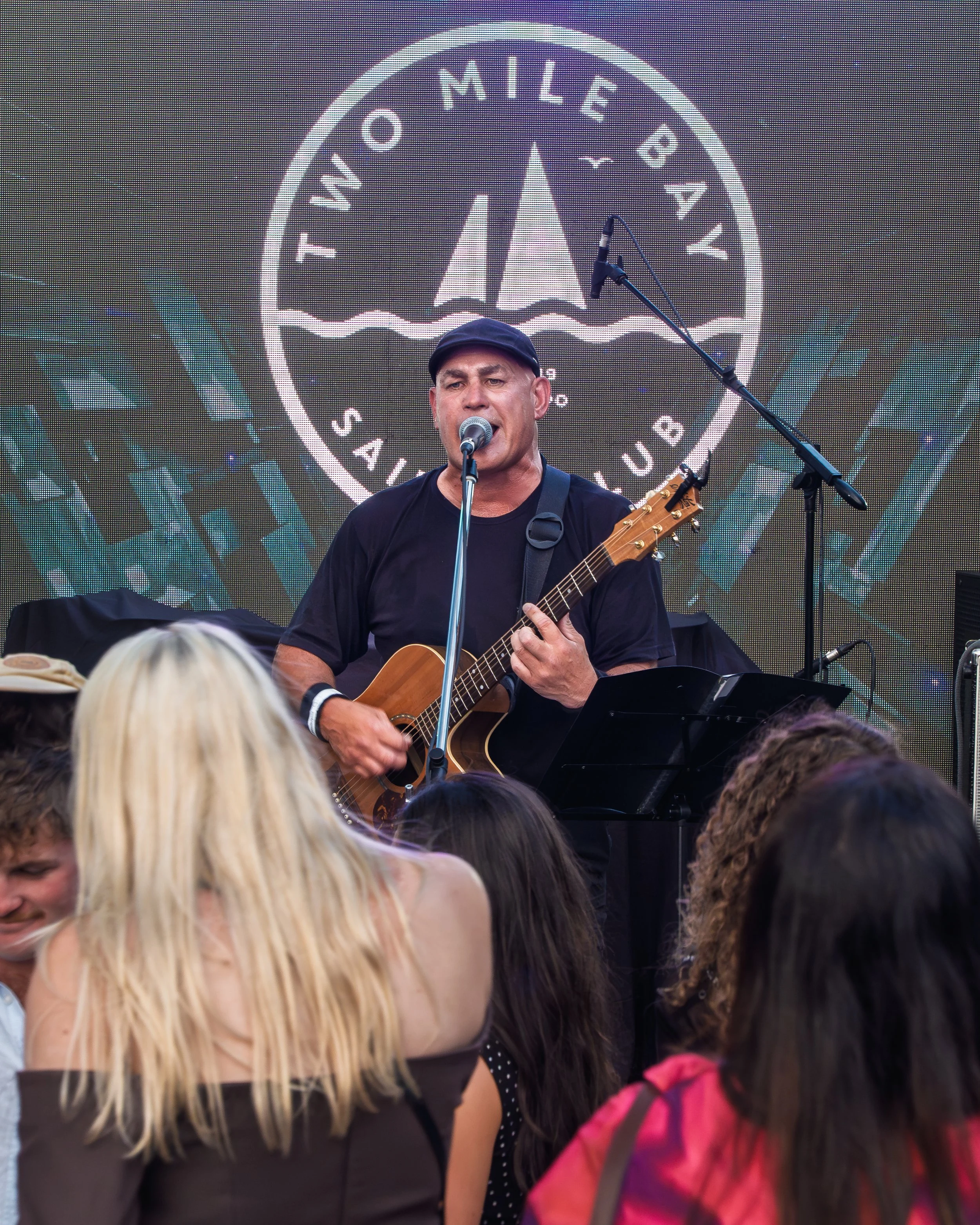 Close-up of a person playing an acoustic guitar, with the text 'LIVE MUSIC' and wavy lines overlayed, and a small text '2MILE2GOOD' in the bottom right corner.