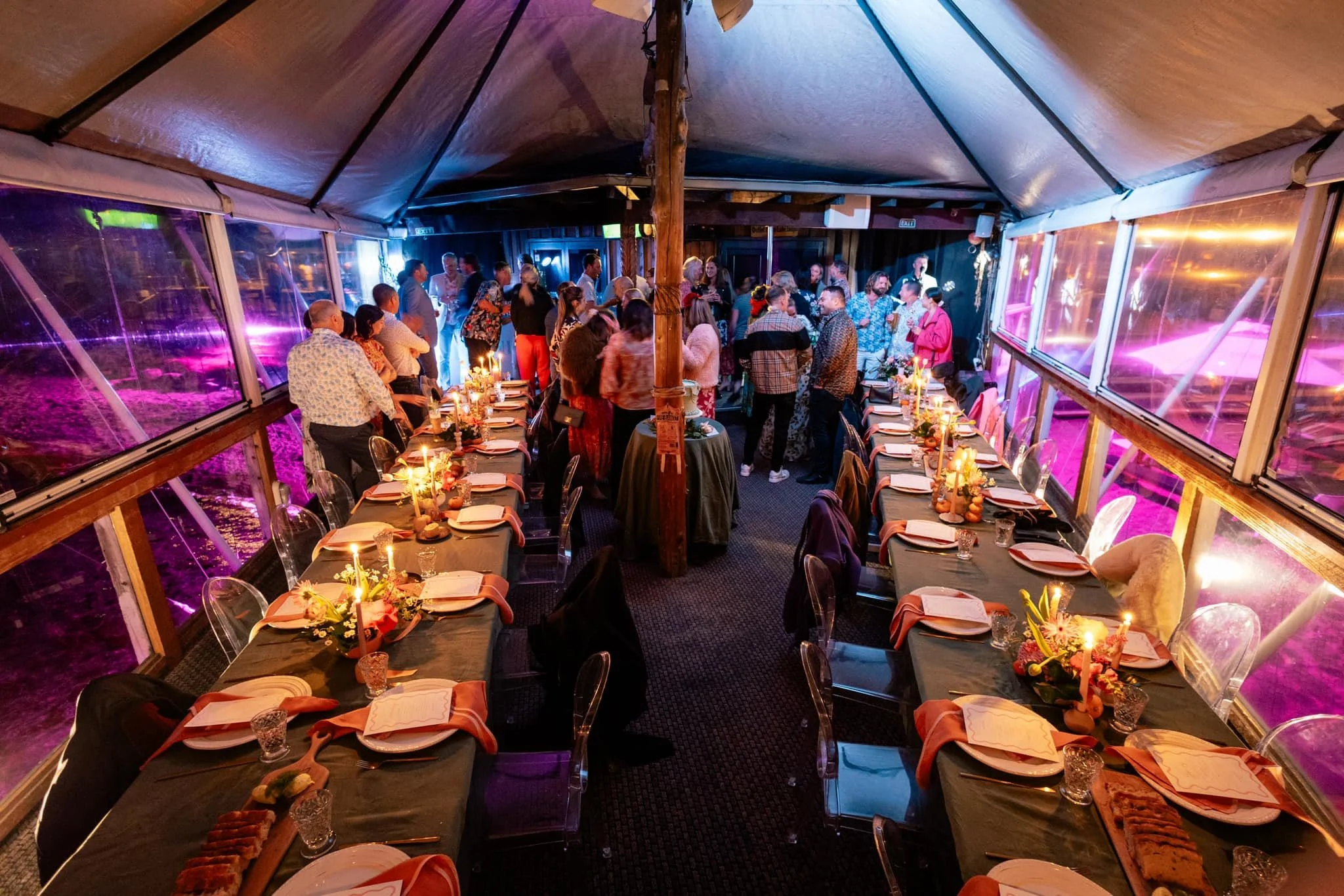 A large indoor banquet hall decorated for a celebration, with tables set with flowers, candles, dinnerware, and glasses, and guests socializing and dancing under a tent-like ceiling with large windows showing colorful lights outside.