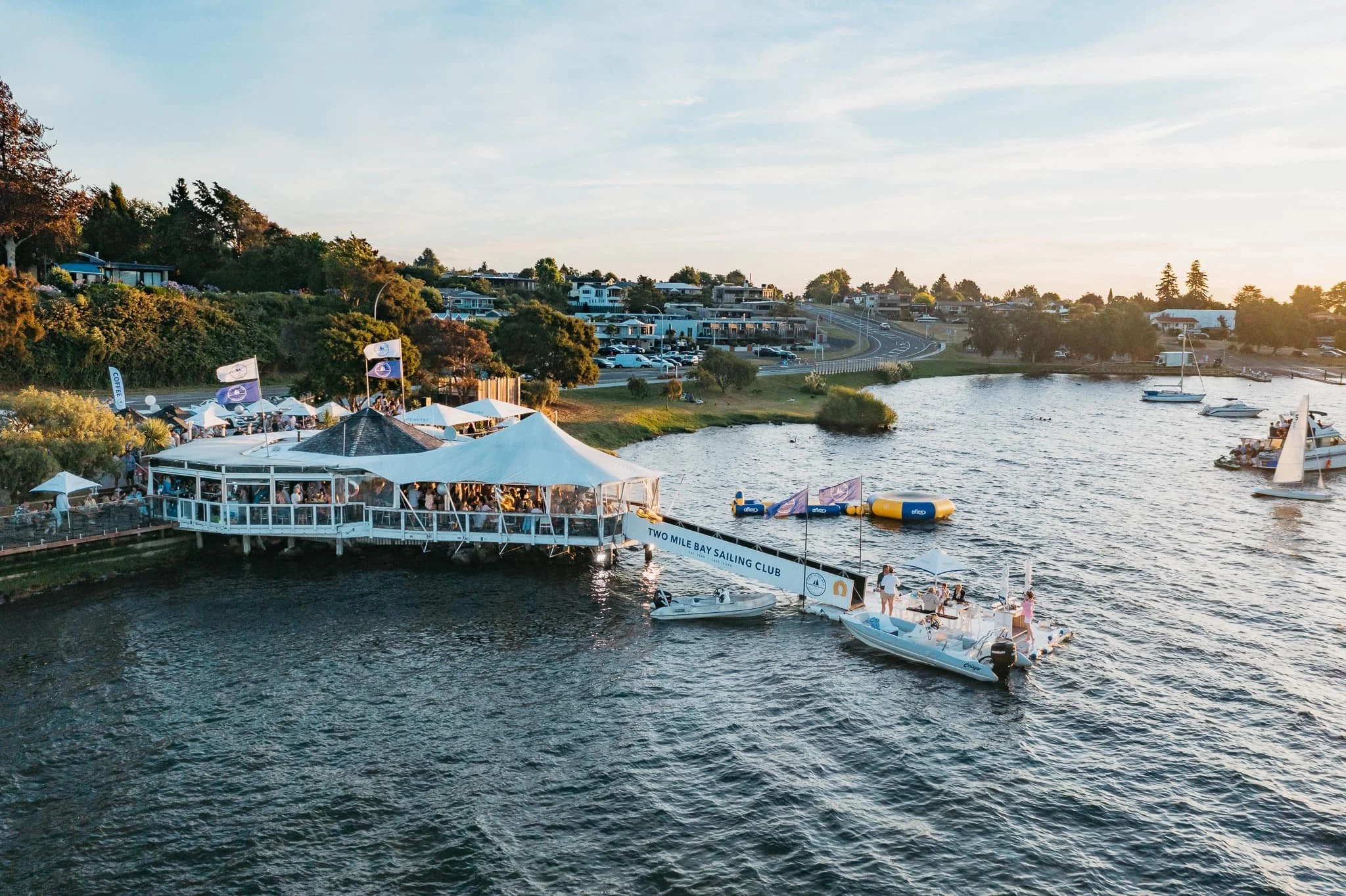 A lakeside scene at sunset showing a marina with boats, a floating dock with people, and a lakeside restaurant with tables and flags.