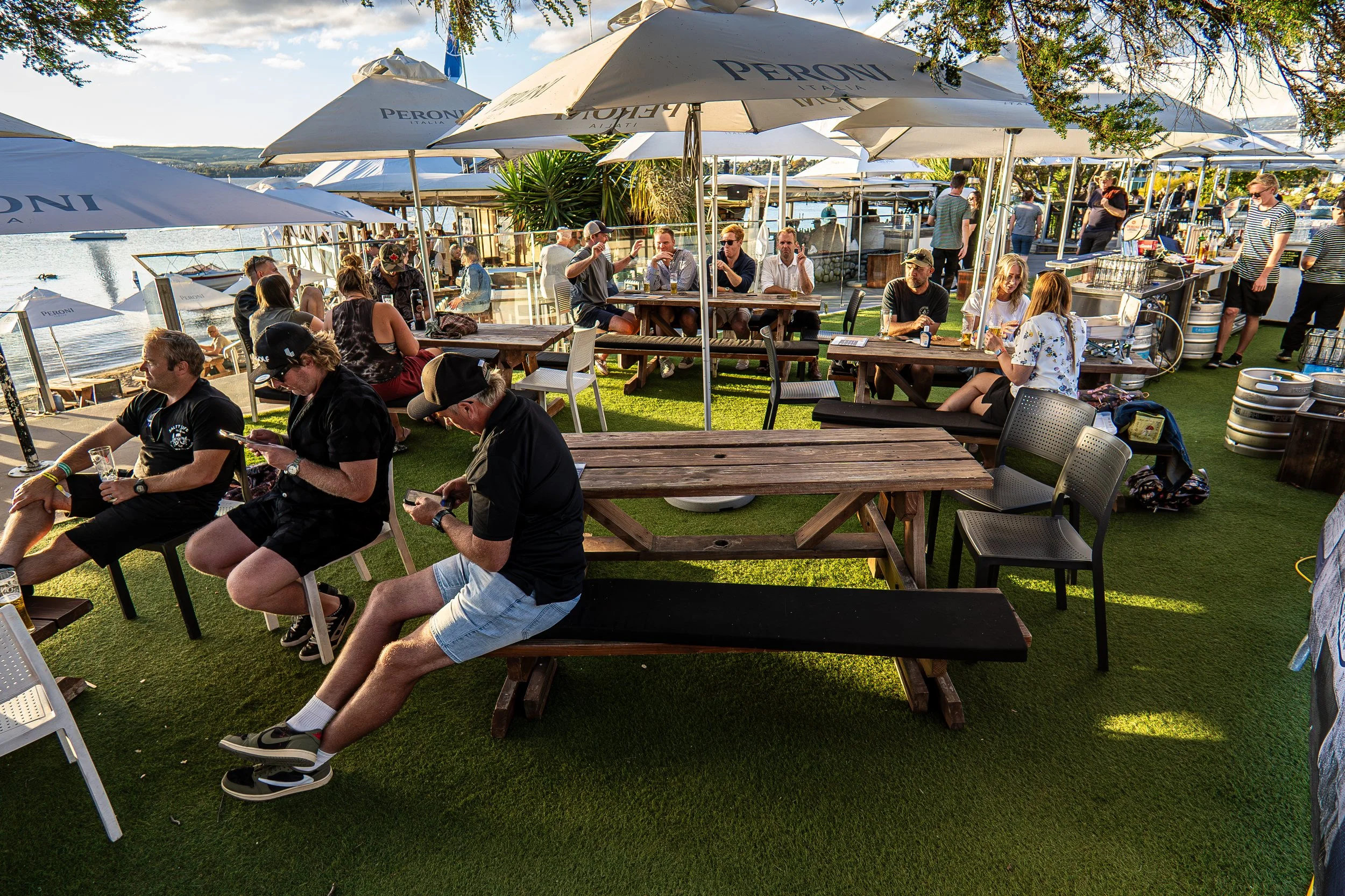 Outdoor café with people sitting at tables under umbrellas near a body of water, some are eating, drinking, and using phones on a sunny day.