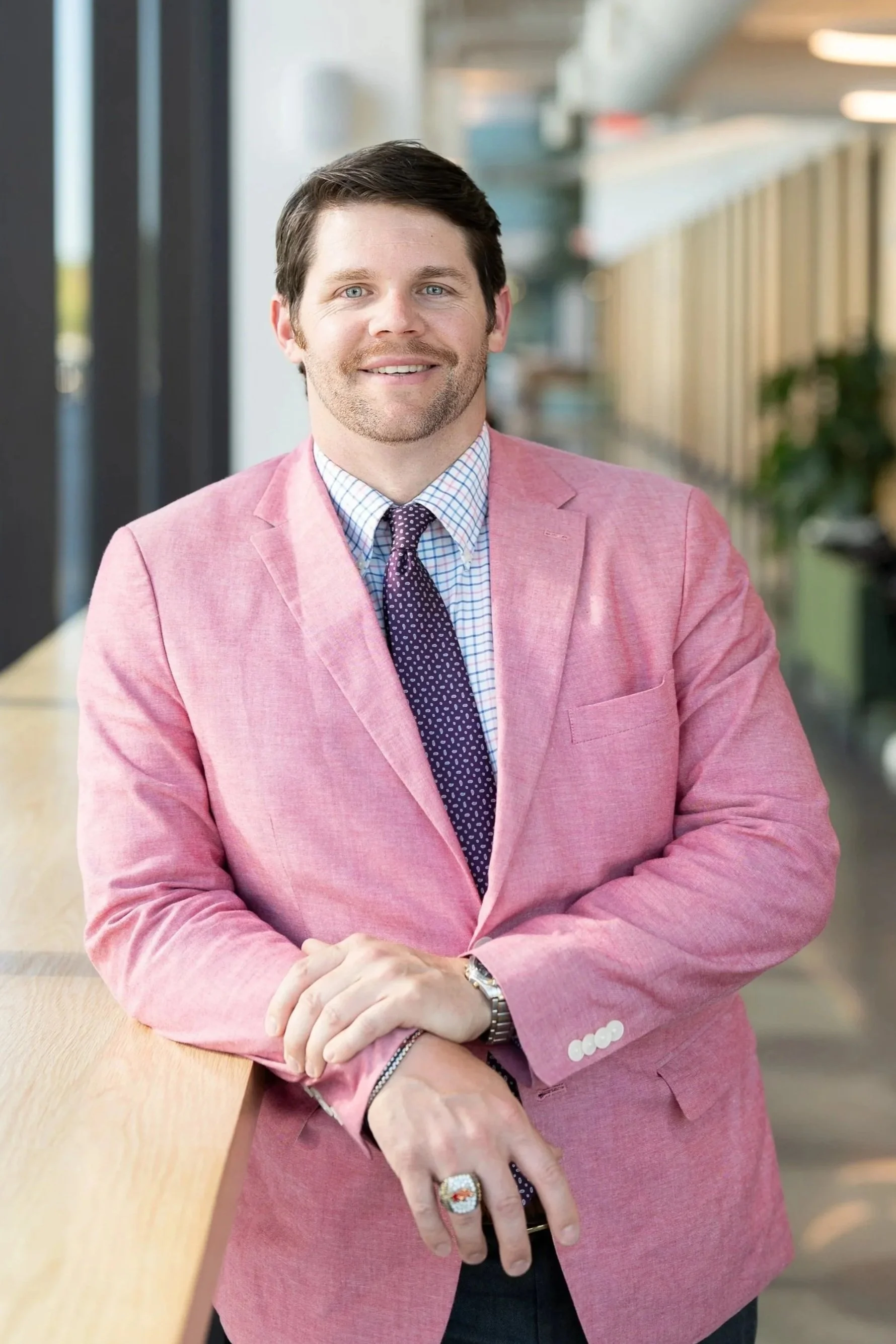 Portrait of a smiling man with brown hair, blue eyes, wearing a pink blazer, checkered shirt, and a dark purple tie. He is standing inside a modern building with large windows and a long corridor.