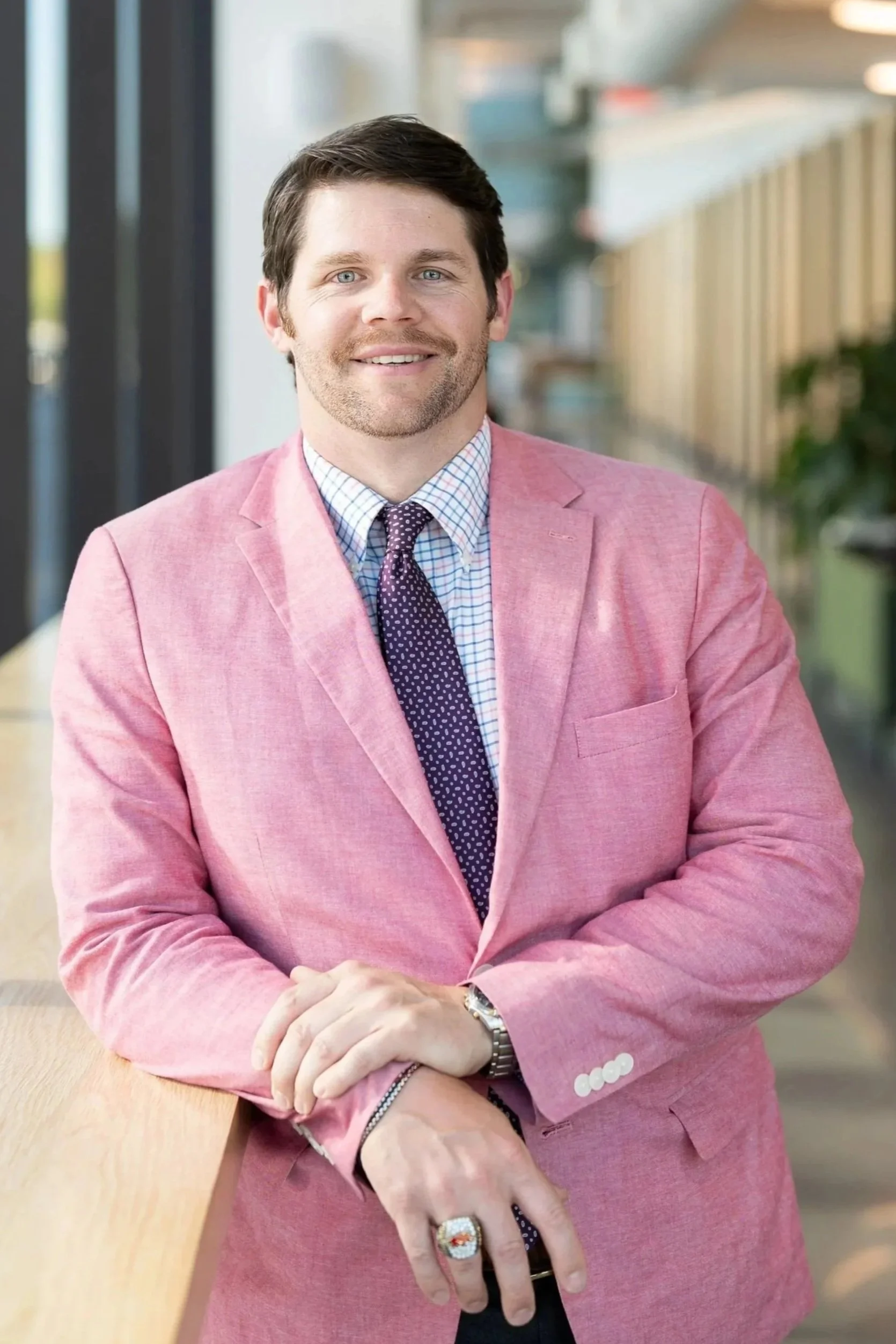 A man with dark hair, blue eyes, and a beard, wearing a pink blazer, checkered shirt, and purple dotted tie, standing indoors with a smile.