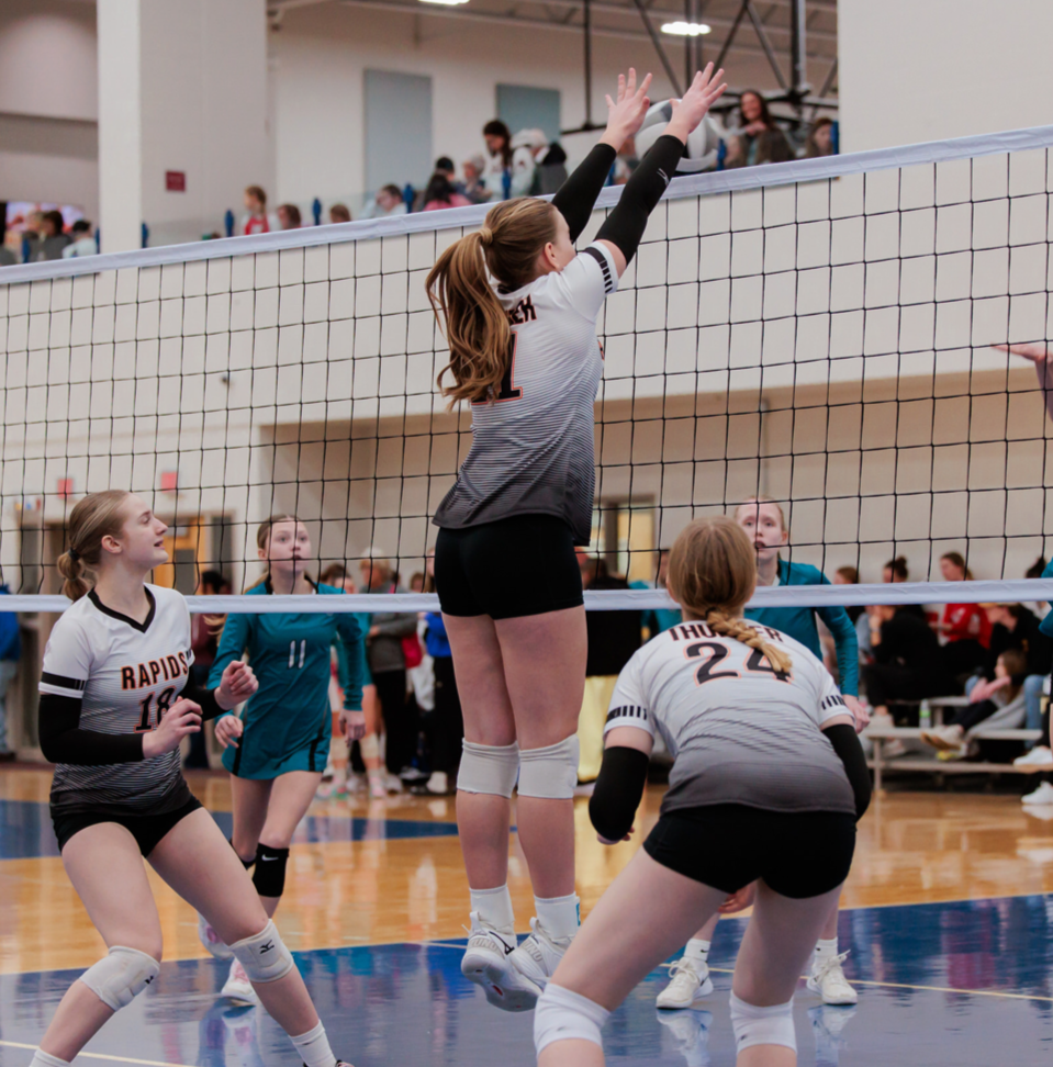 A girls' volleyball game with players jumping at the net in an indoor gymnasium. One player is blocking, others are preparing to hit the ball.