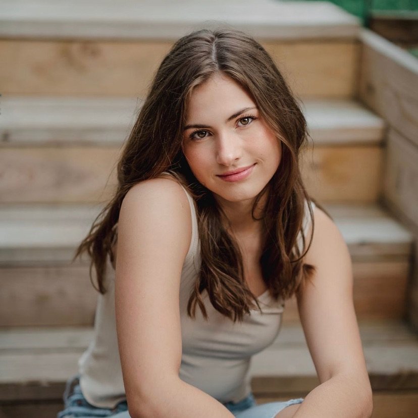 A young woman with long brown hair sitting on wooden stairs, smiling softly at the camera.