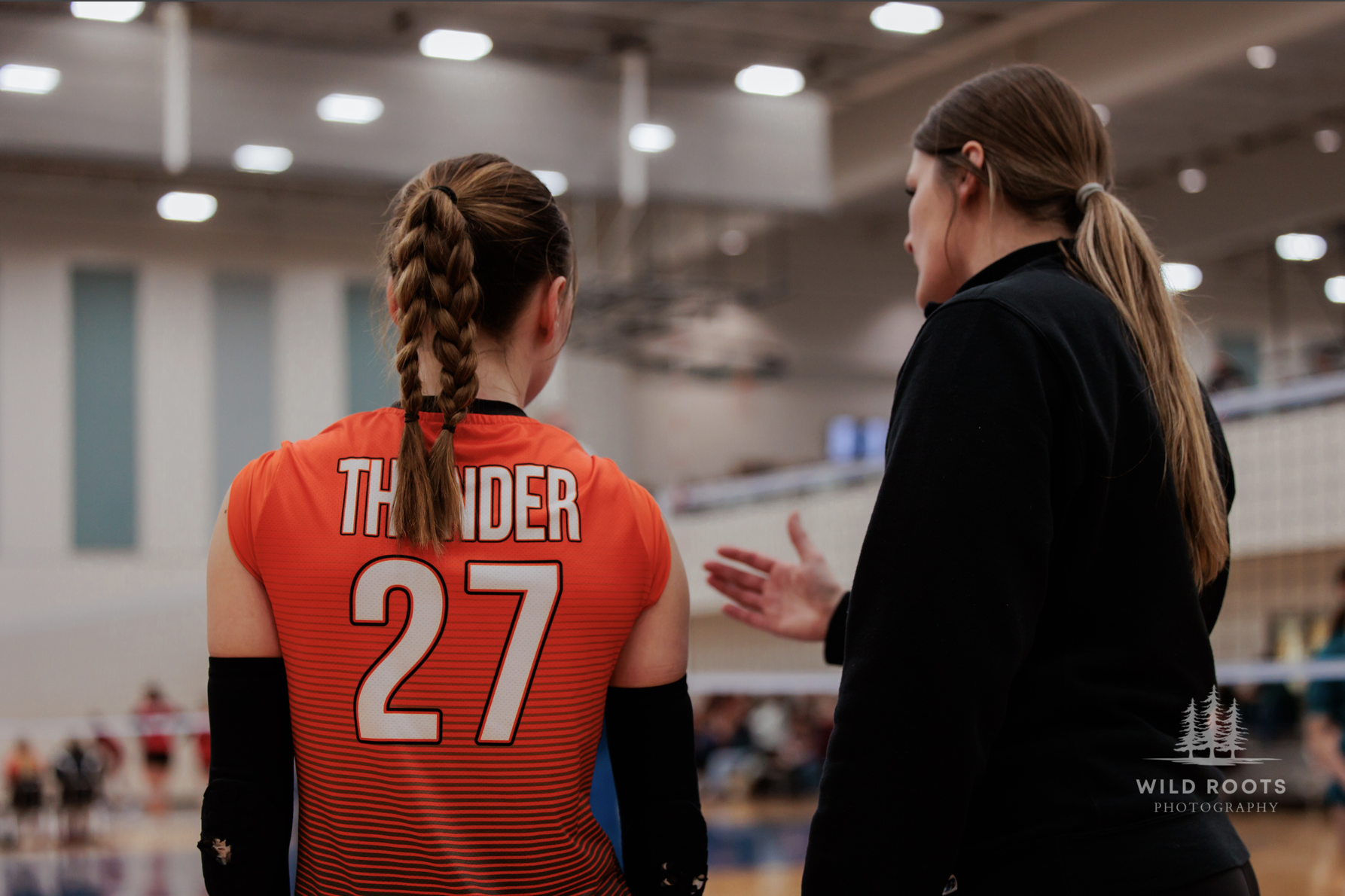A female volleyball player wearing a red jersey with the name 'THUNDER' and the number 27 on her back, talking to a female coach or teammate in a black jacket inside an indoor gymnasium.