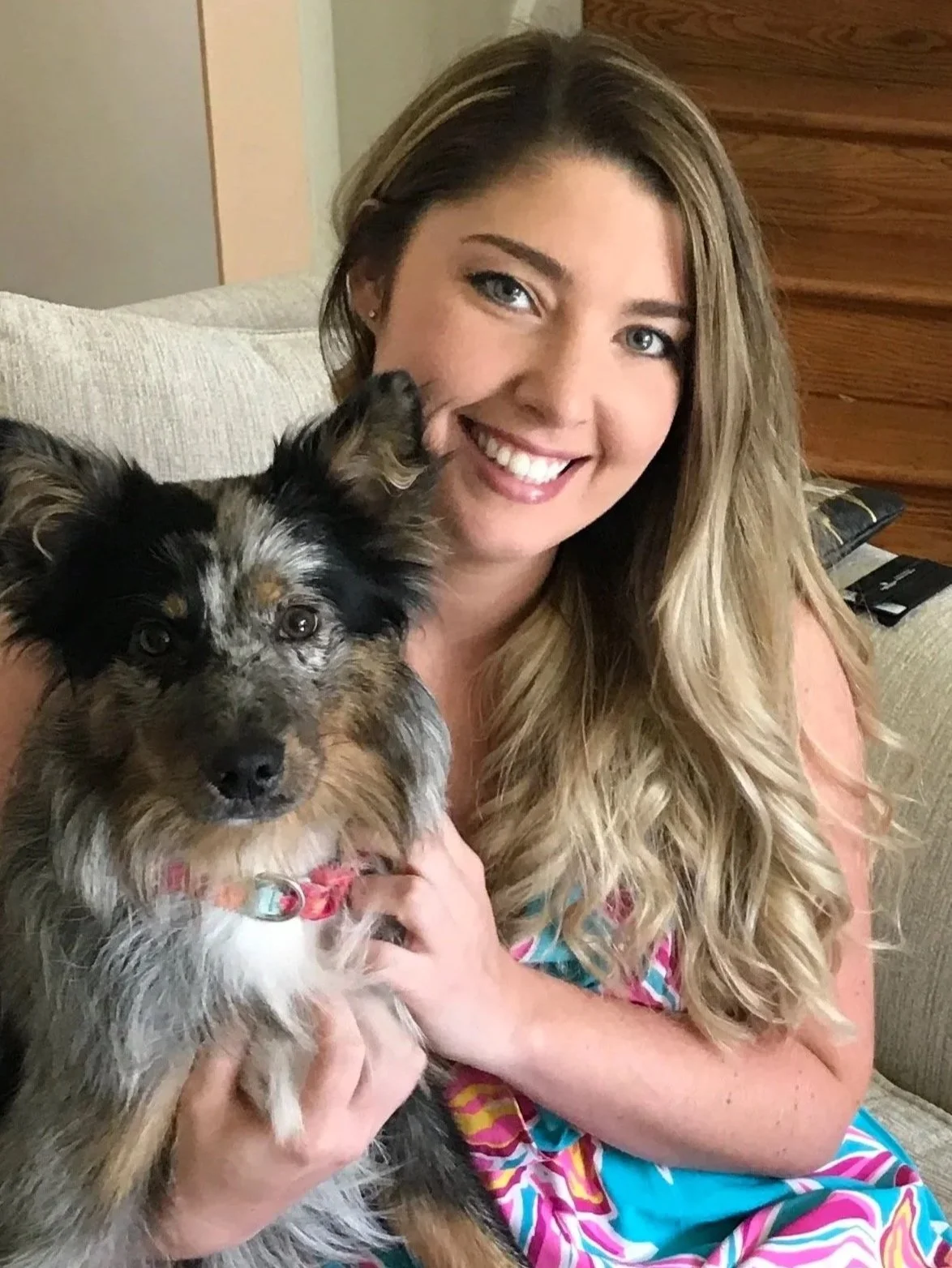 A woman with long, wavy blonde hair smiling and holding a small, multicolored Australian Shepherd puppy inside a living room.