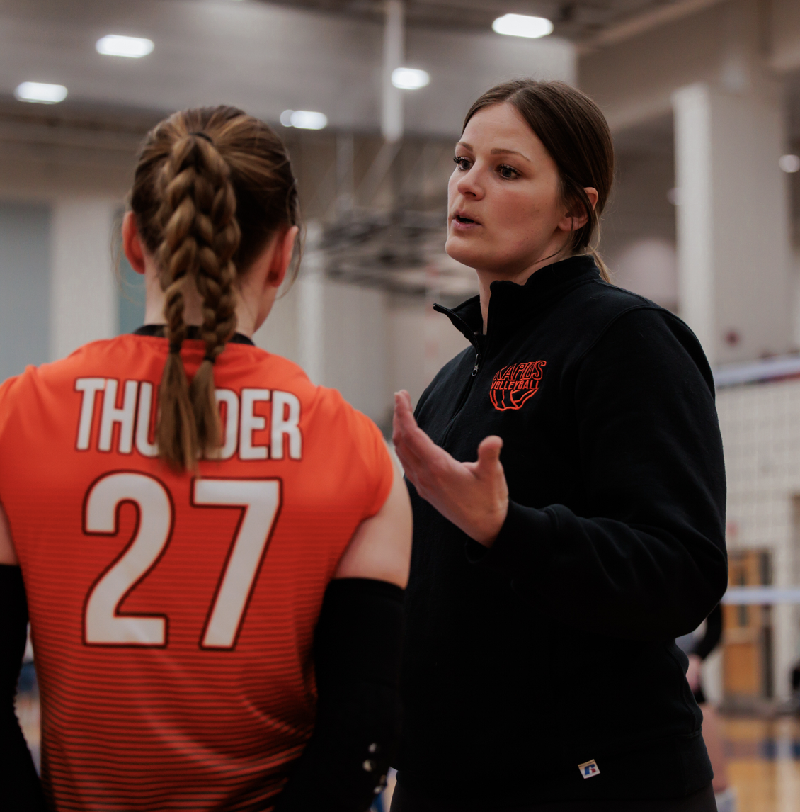 A volleyball coach talking to a player in a gymnasium, with the player wearing an orange jersey with 'THUNDER' and the number 27 on the back.