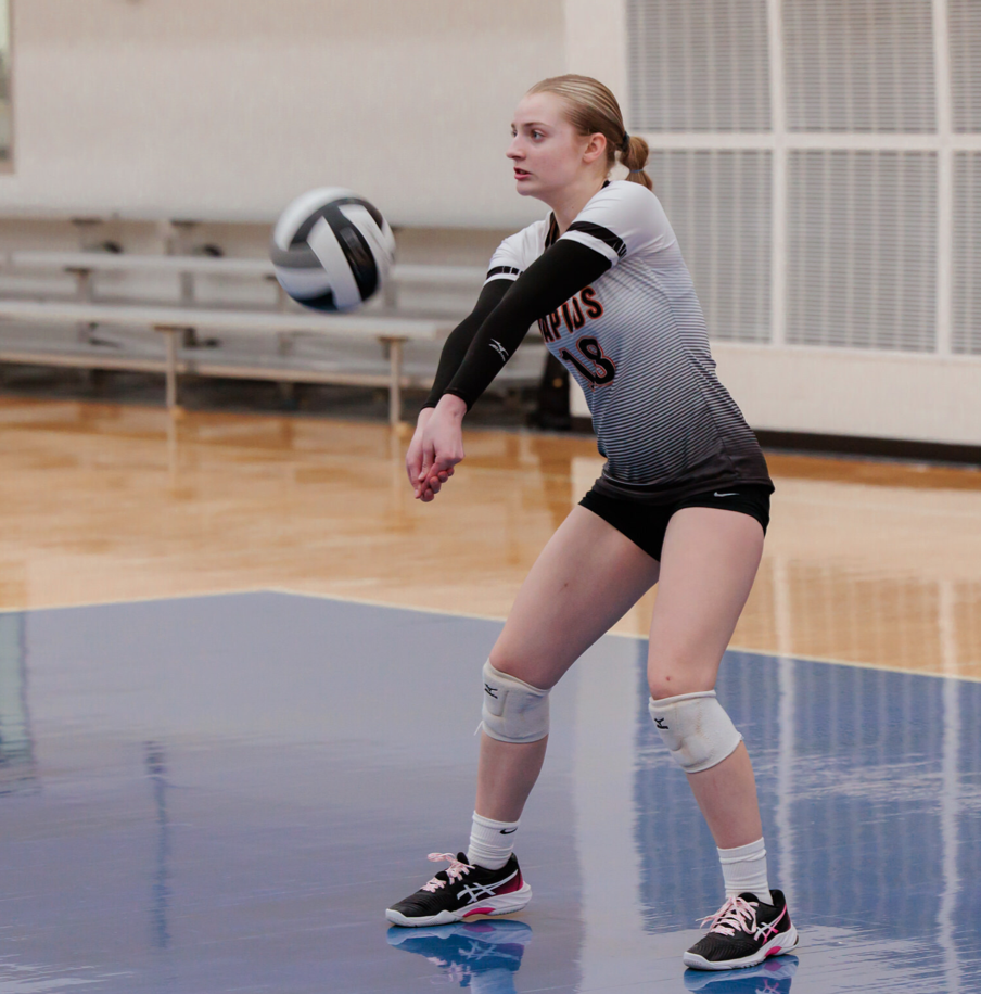 A female volleyball player in a white and black uniform with the number 18, wearing knee pads and sneakers, is preparing to pass a volleyball on an indoor court.