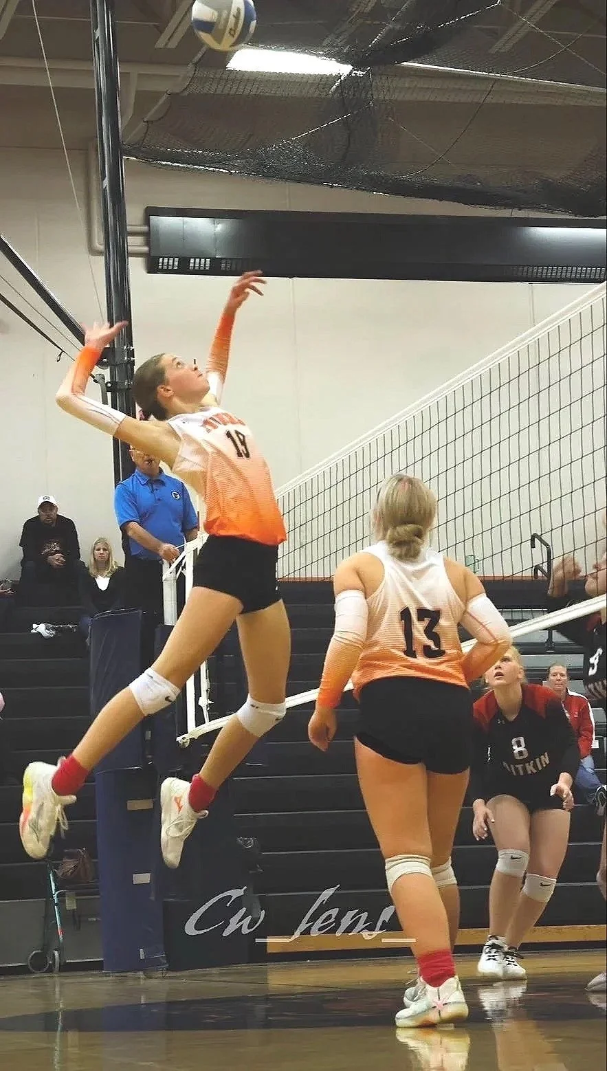 A volleyball player in mid-air hitting the ball over the net while teammates and an opponent watch.
