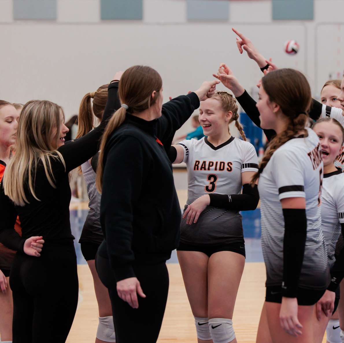 Girls volleyball team celebrating with coach during game in indoor gym.