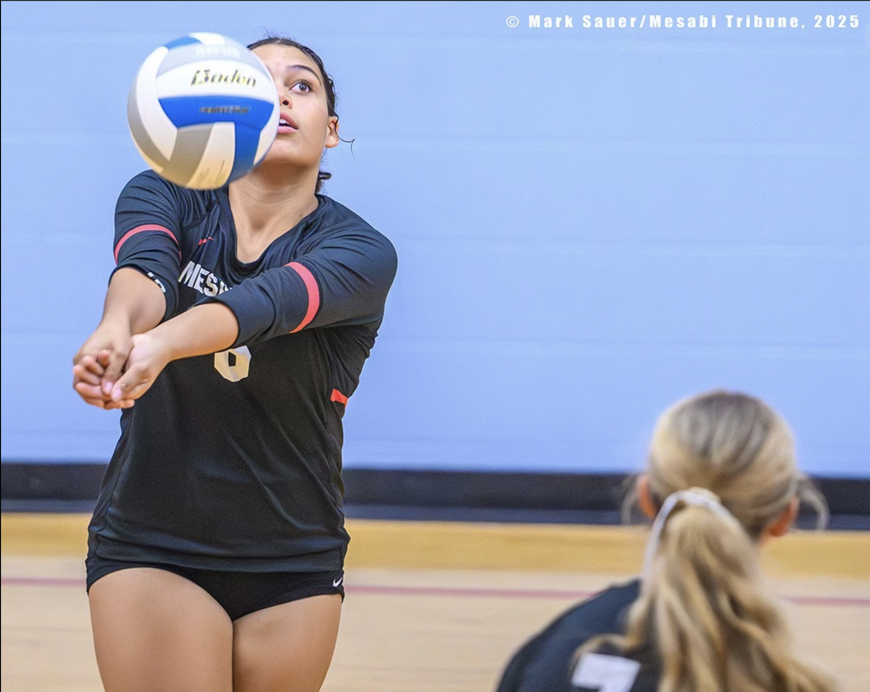 A female volleyball player is hitting a volleyball in an indoor gym, with another player seated in front watching.