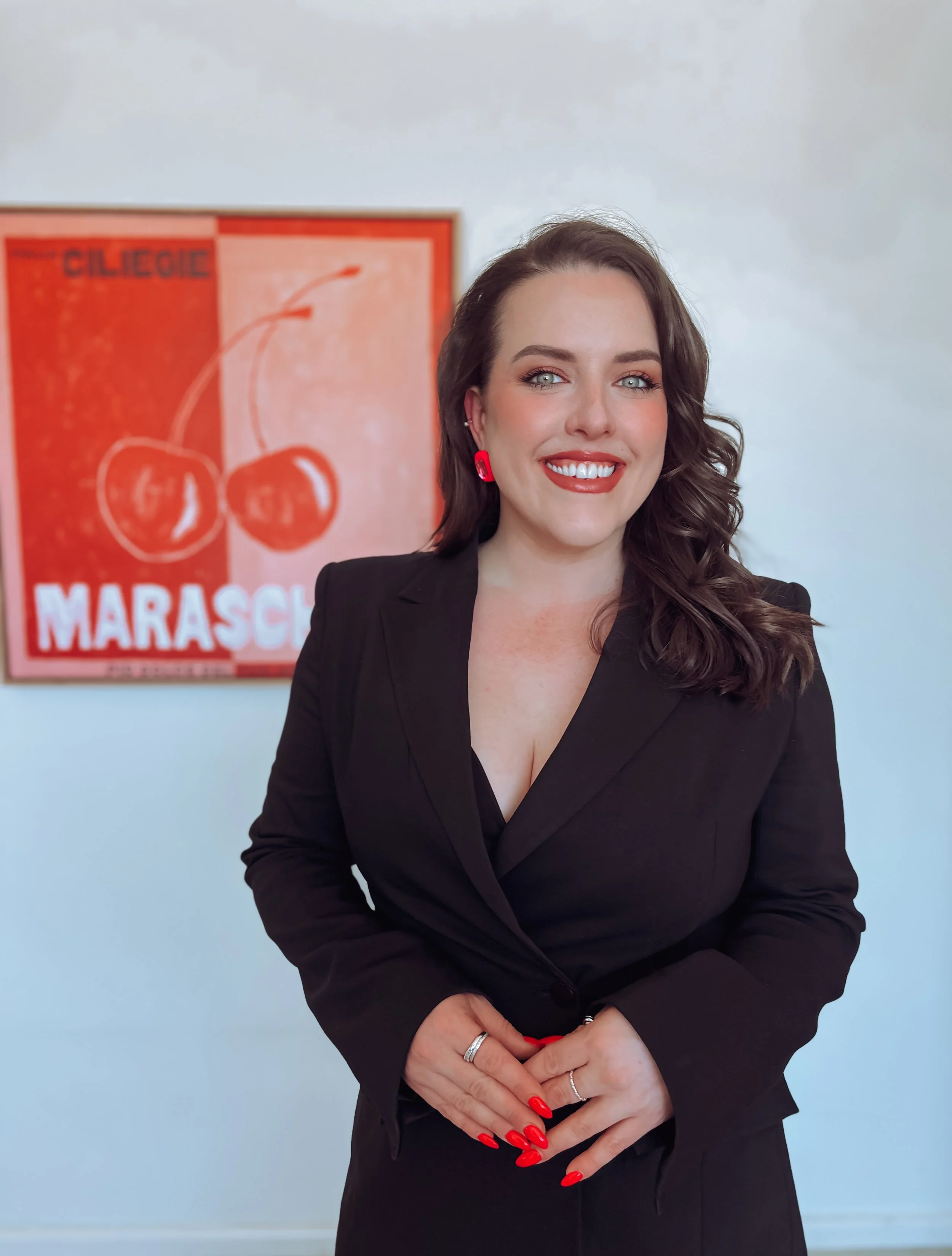 A woman with dark wavy hair, wearing a black blazer, red earrings, and red nails, smiling and standing in front of a cherry-themed poster.