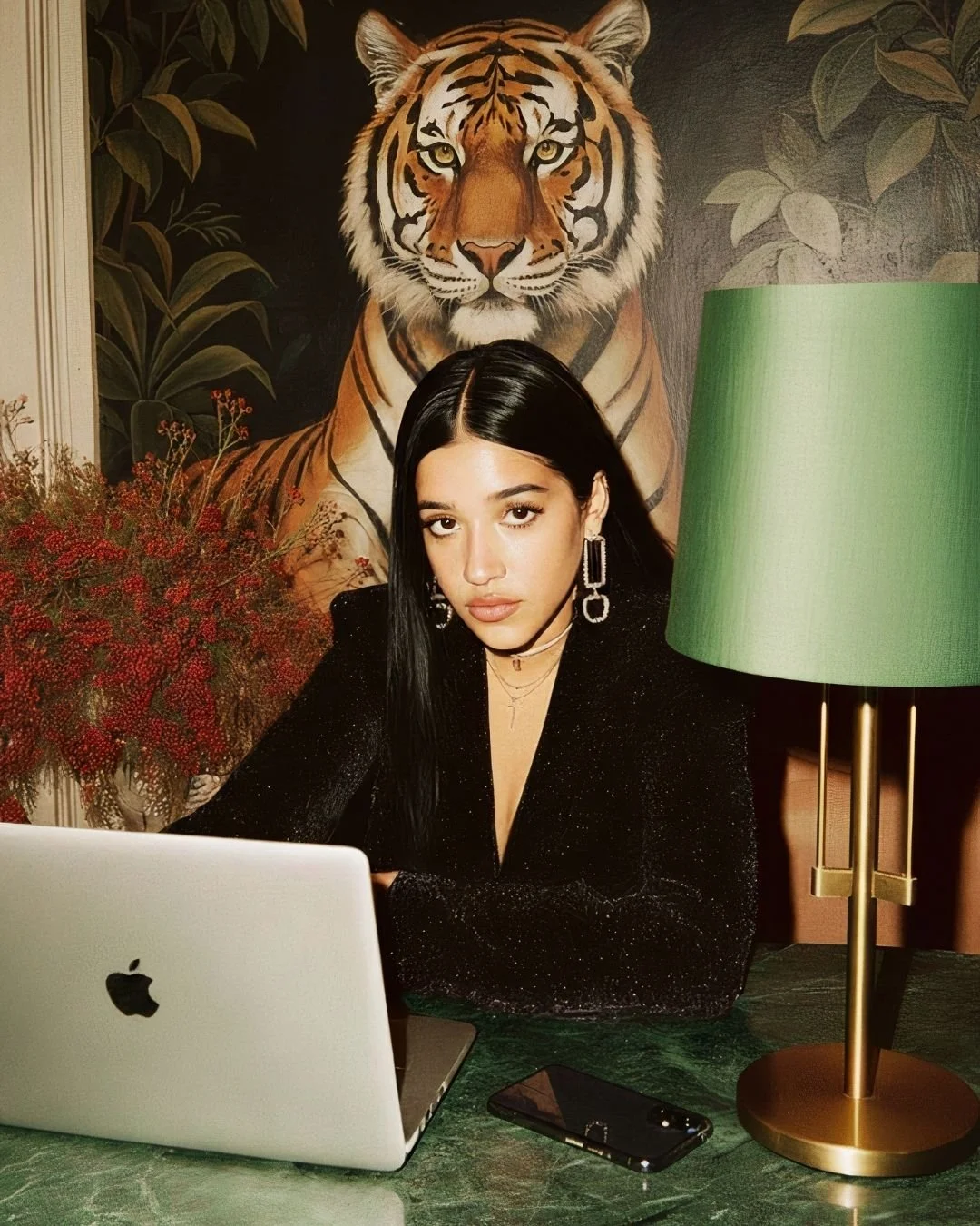 A woman with long black hair and hoop earrings sitting at a green table with a silver laptop and a smartphone, in front of a tiger painting and a plant with red flowers, and a green lampshade on a brass lamp.