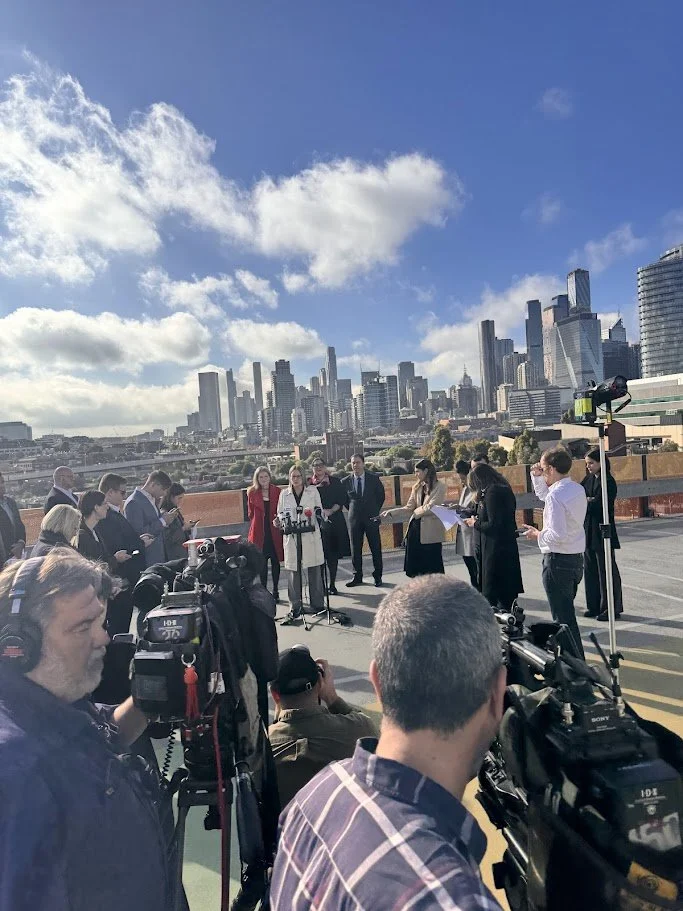 A news conference or press event taking place outdoors on a rooftop parking lot with a city skyline in the background. Several people, including journalists and camera operators, are gathered around speakers at the podium.