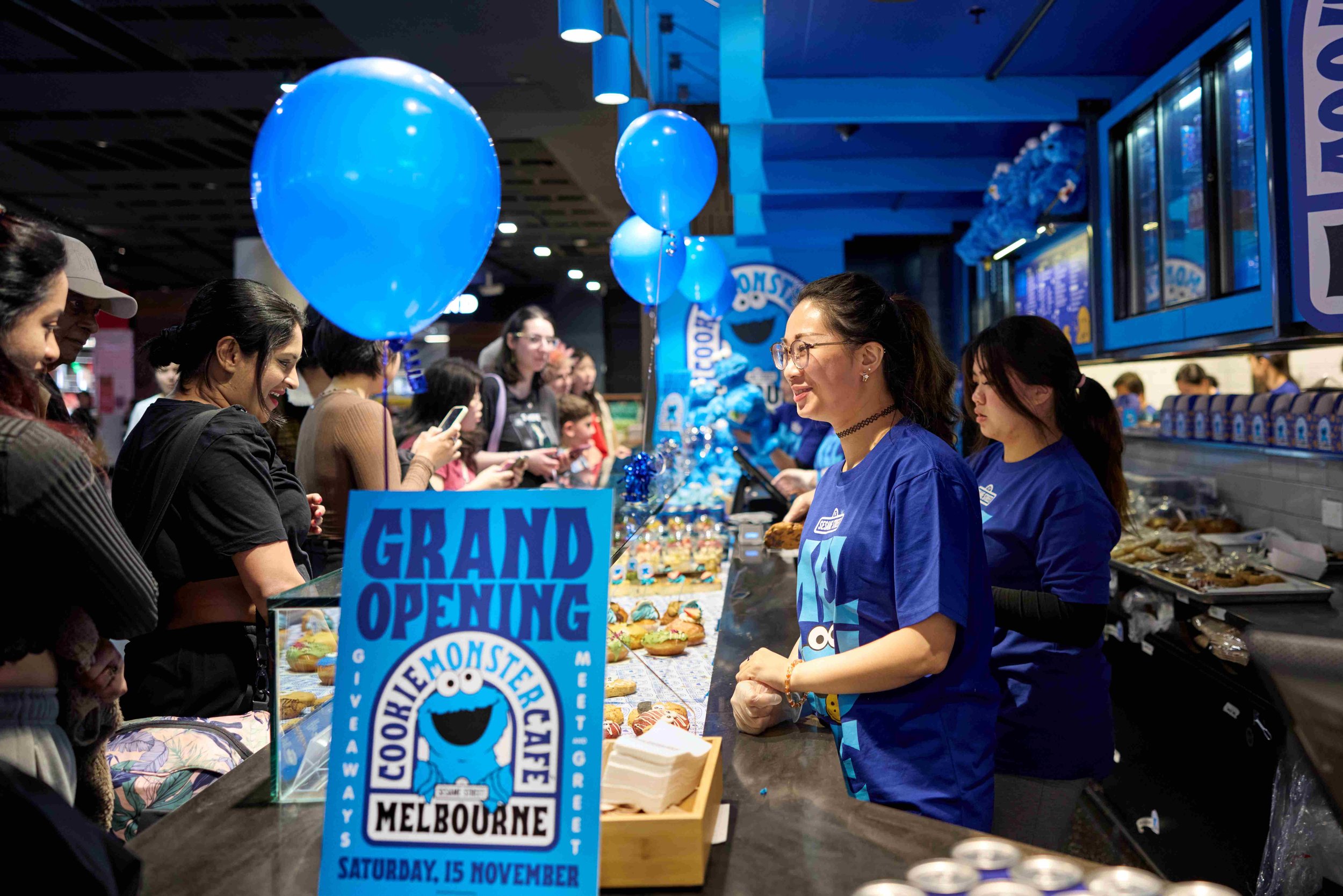 People waiting in line at a Japanese bakery's grand opening event with a promotional poster, blue balloons, and bakery items on the counter.