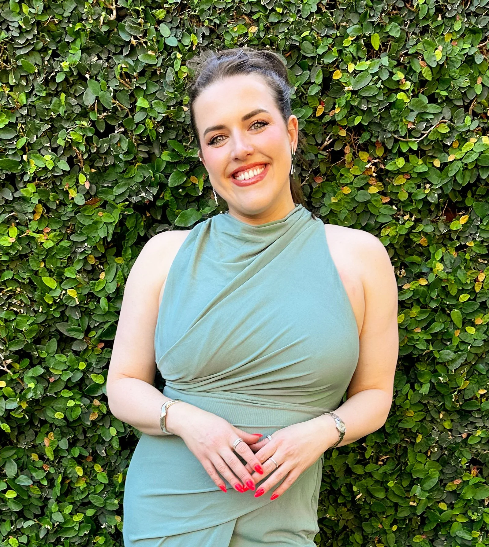 A woman with dark hair in a ponytail, wearing a light green sleeveless dress, smiling, standing in front of a green leafy hedge.