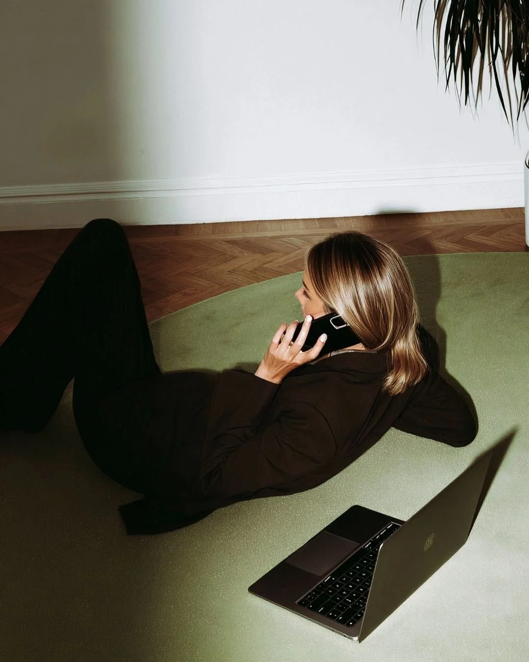 Woman lying on a green rug talking on a black landline phone with an open laptop nearby.