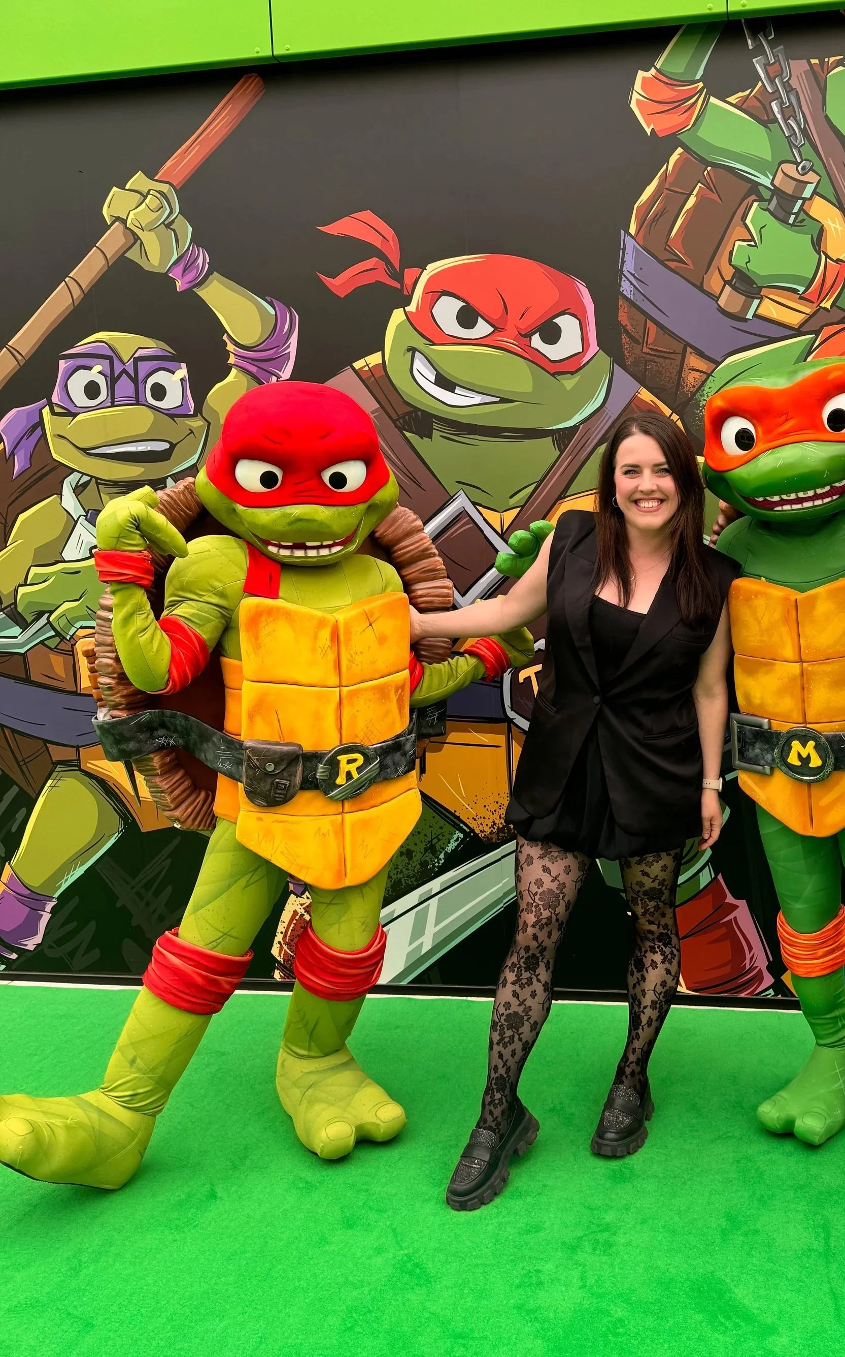 A woman posing with life-sized Teenage Mutant Ninja Turtles characters at the launch event for the exhibition. The background features a large Teenage Mutant Ninja Turtles graphic poster.