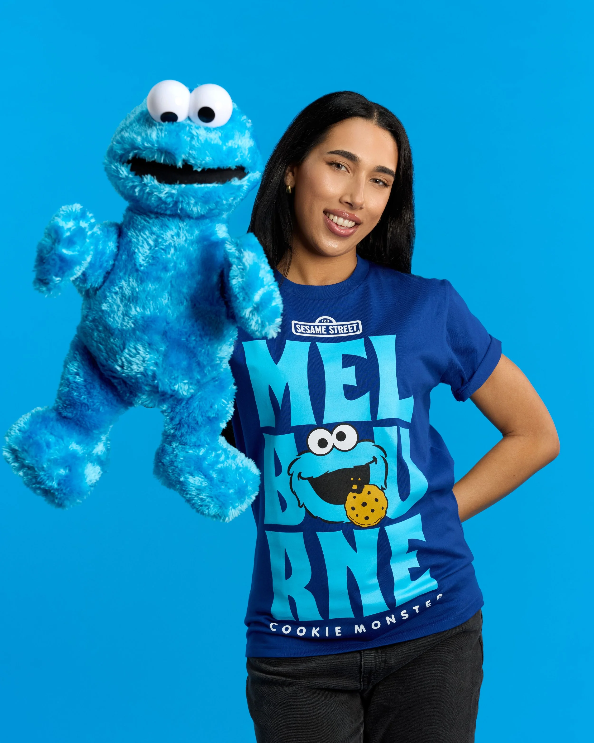 Woman in blue Sesame Street T-shirt holding plush Cookie Monster puppet against a blue background.