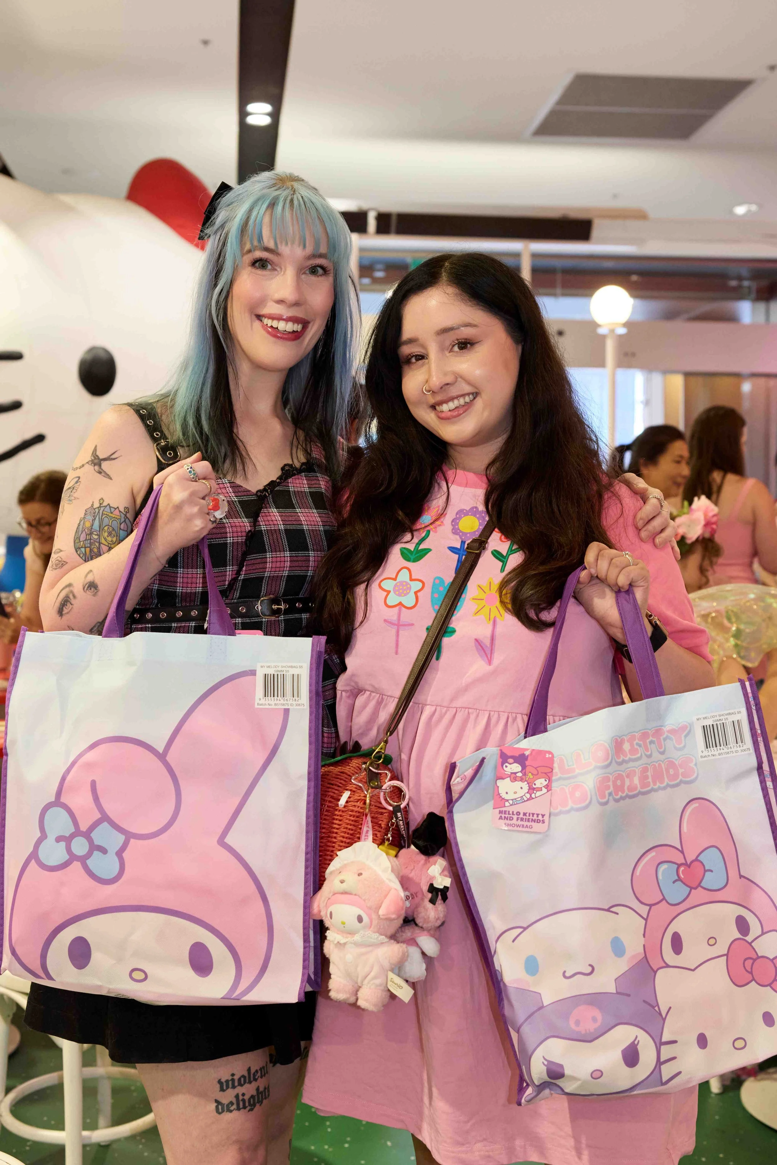 Two women smiling and holding Hello Kitty themed shopping bags at a party or event, with other people and pink decor in the background.