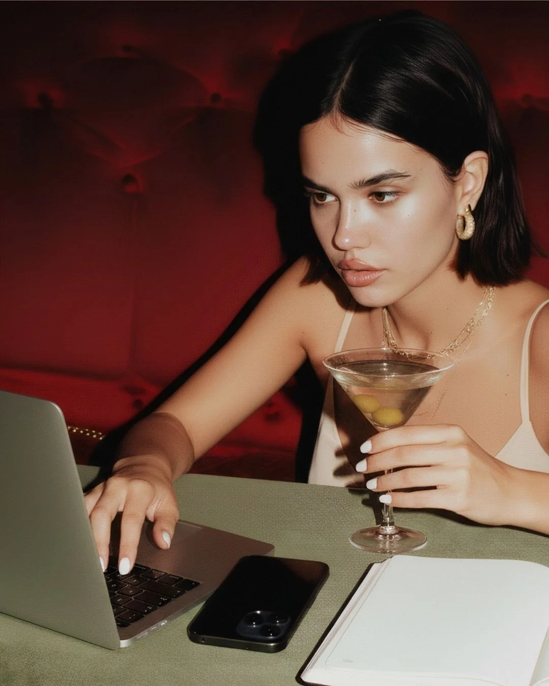 A young woman with dark hair, wearing jewelry, using a laptop at a table with a smartphone and a notebook, holding a martini glass with olives, sitting in a dimly lit setting.