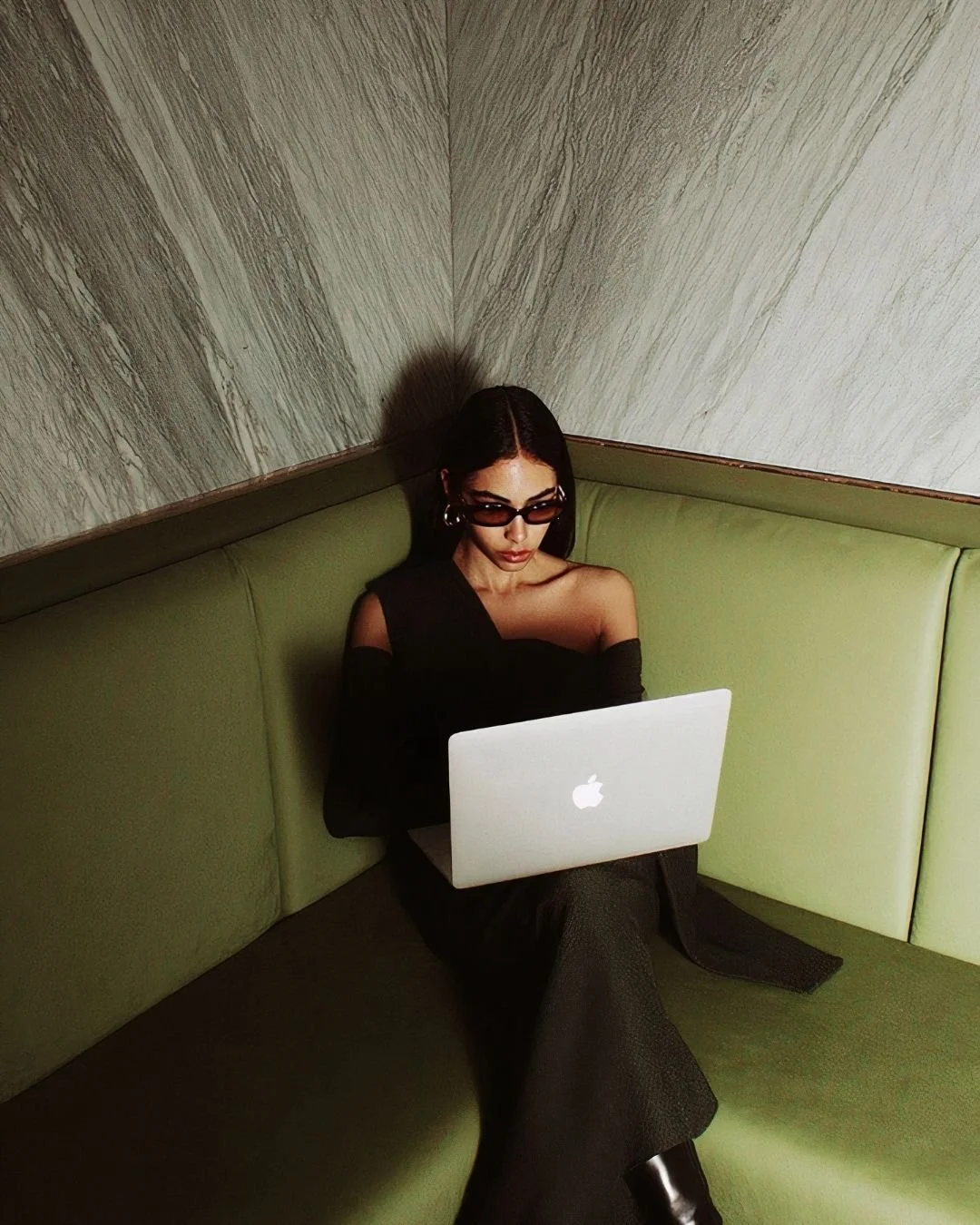 A woman with dark hair, wearing black sunglasses and a black off-shoulder top, sitting on a green booth with a laptop in her lap, looking focused.