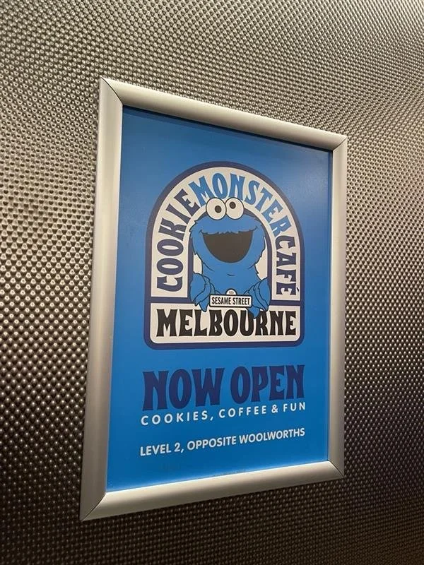 Sign for the Cookie Monster Cafe in Melbourne, featuring Cookie Monster's face, indicating it is open for cookies, coffee, and fun, located on Level 2 across from Woolworths.