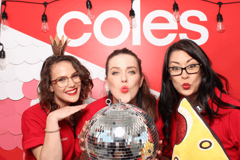 Three women wearing red shirts and glasses posing with a disco ball in front of a red and pink backdrop with 'Coles' logo. One woman is wearing a crown, another is holding a large pizza cutout, and they are all making playful expressions.