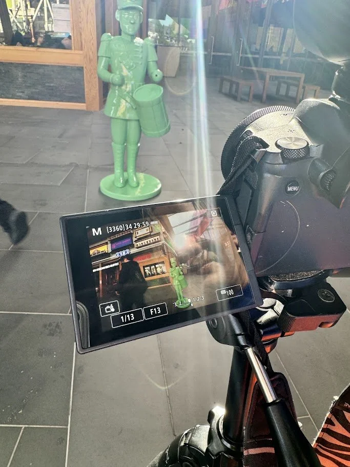 A green toy soldier sculpture playing a drum outside in front of a window, with a camera on a tripod in the foreground capturing the scene.