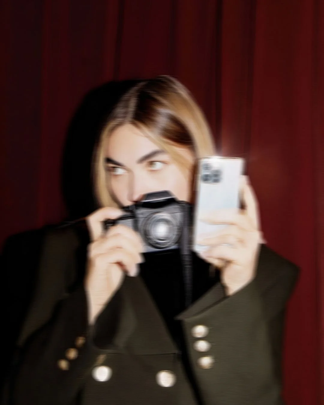 A person with two-toned hair, holding a camera and a smartphone, looks to the side, standing in front of a dark red curtain.