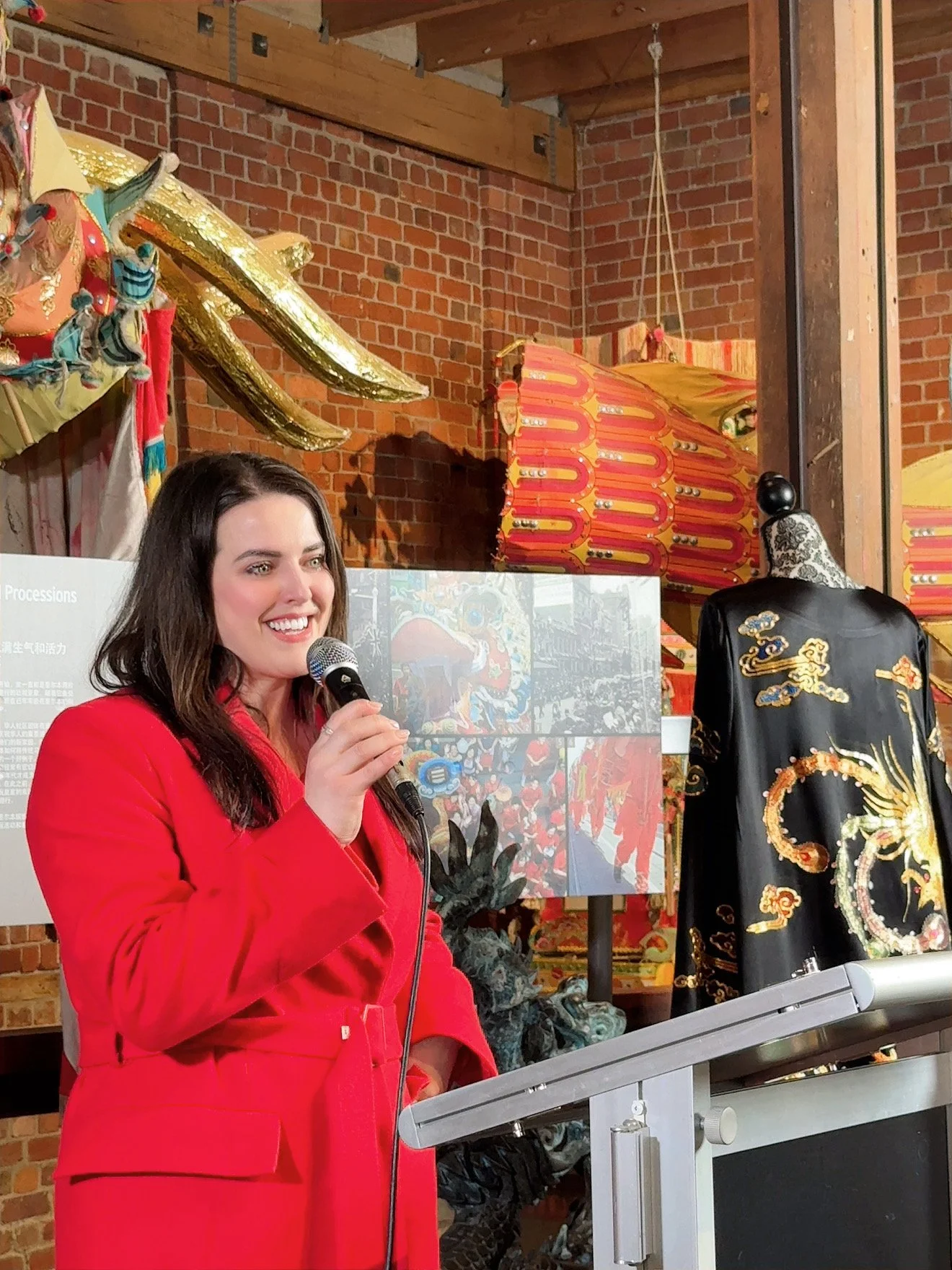 A woman in a red blazer speaking into a microphone at an indoor event, with traditional Chinese costumes and decorations displayed behind her.