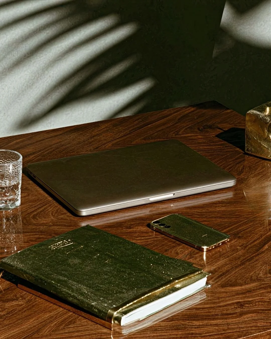 A wooden desk with a closed laptop, a glass of water, a gold-colored notebook, a smartphone, and a gold-colored decorative object, with shadow of plant leaves cast on the wall in the background.