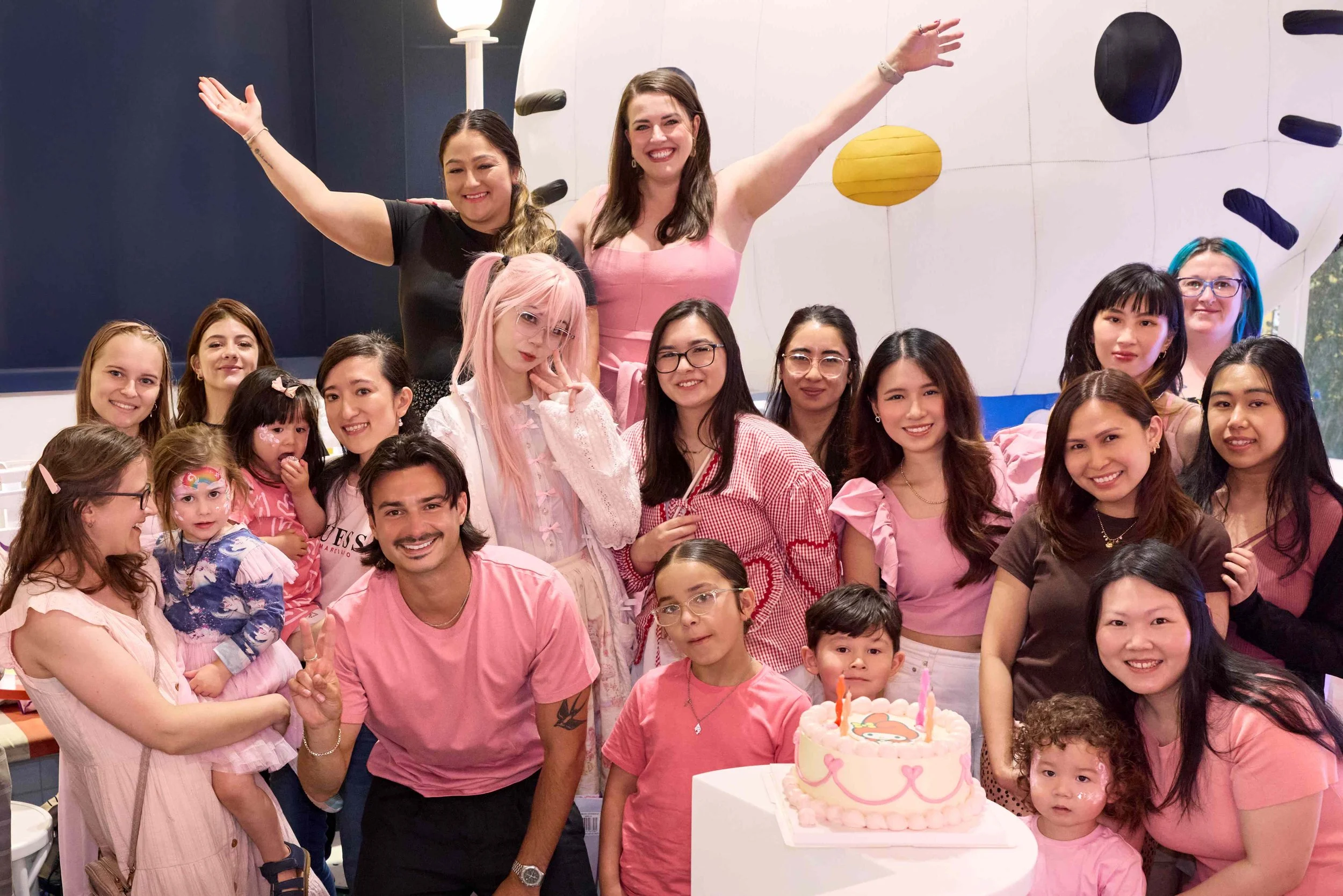 Group of people celebrating a birthday party, with a birthday cake on a table, indoors with a climbing wall in the background.