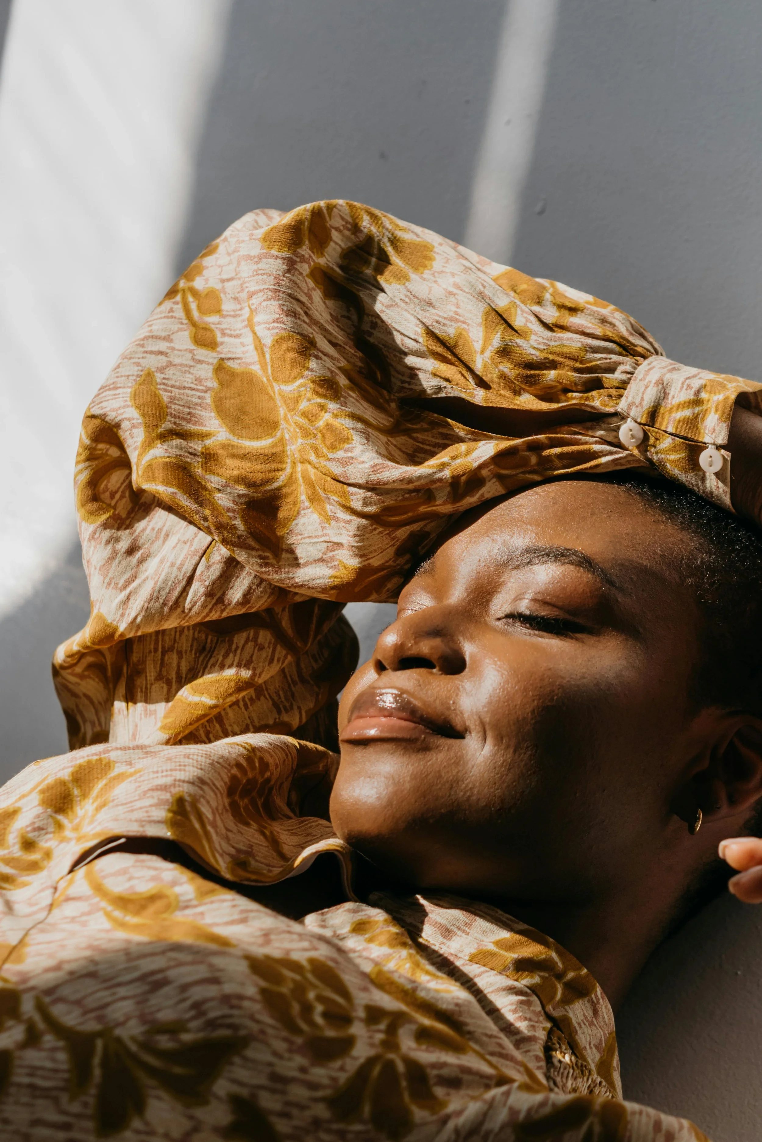 Black woman resting an arm on her forehead with eyes closed and a slight smile, wearing a yellow floral blouse