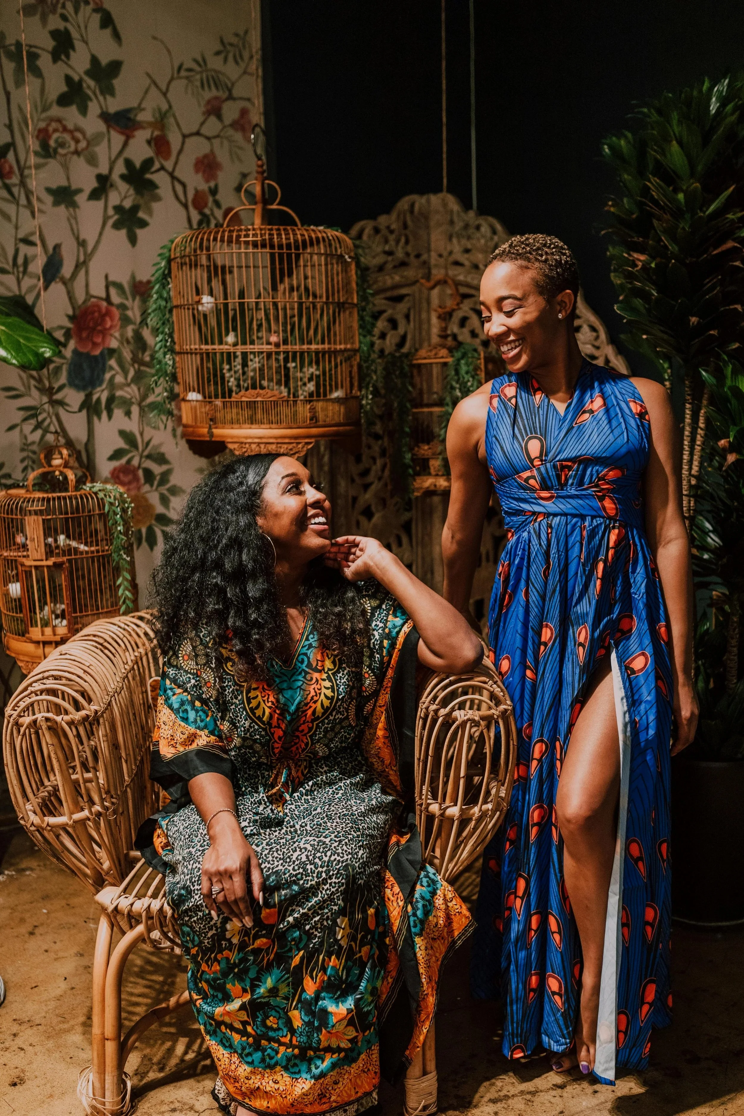 Two Black women smiling at each other — the woman on the left seated in a patterned dress, and the woman on the right with short hair smiling down while wearing a blue flowy dress