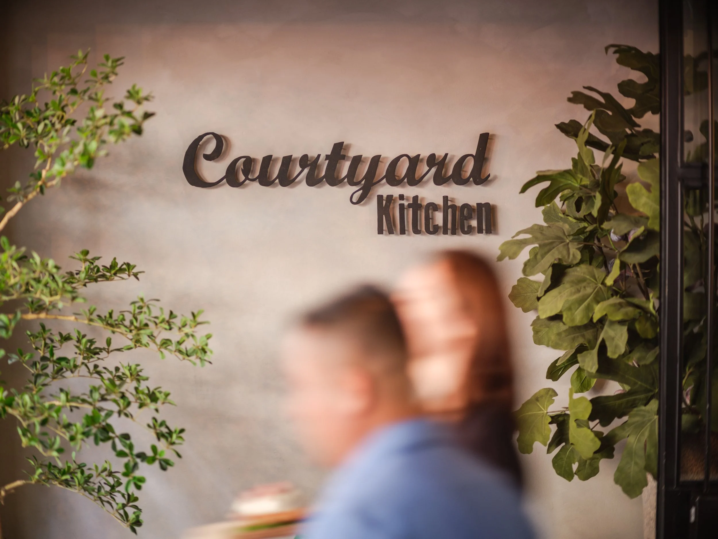 Interior of a kitchen with a sign that reads 'Courtyard Kitchen' on the wall, surrounded by green plants and two blurred people in the foreground.