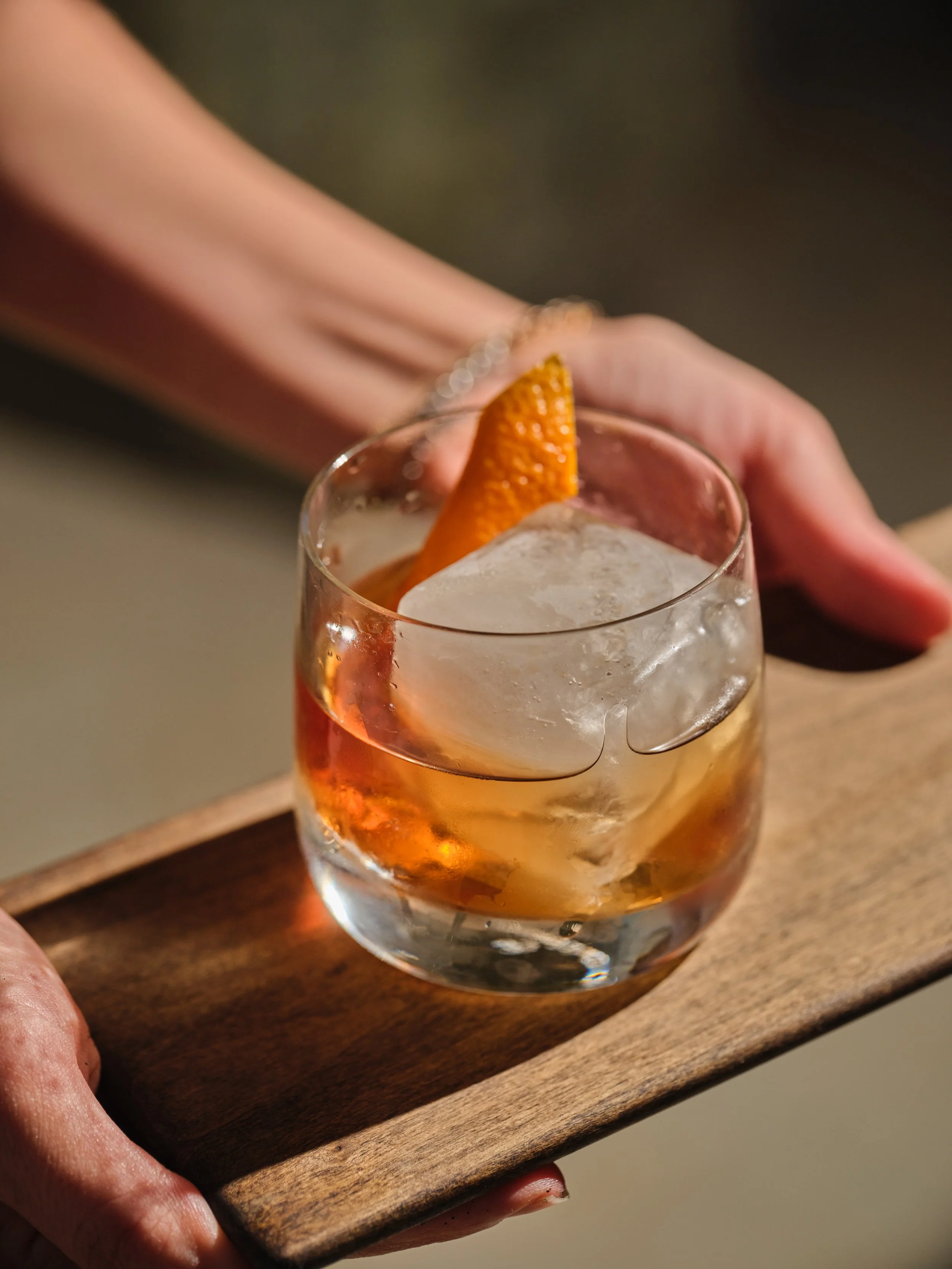 A glass of whiskey garnished with an orange slice, served on a wooden tray.