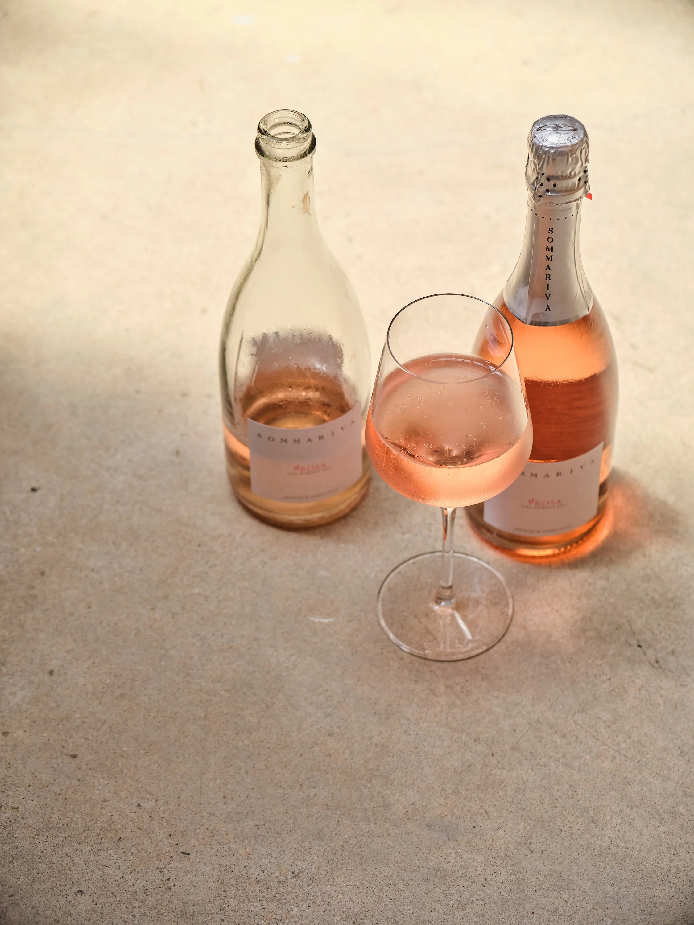A bottle of rosé wine, a partially filled wine bottle with a foil seal, and a wine glass with rosé wine, all on a beige surface.