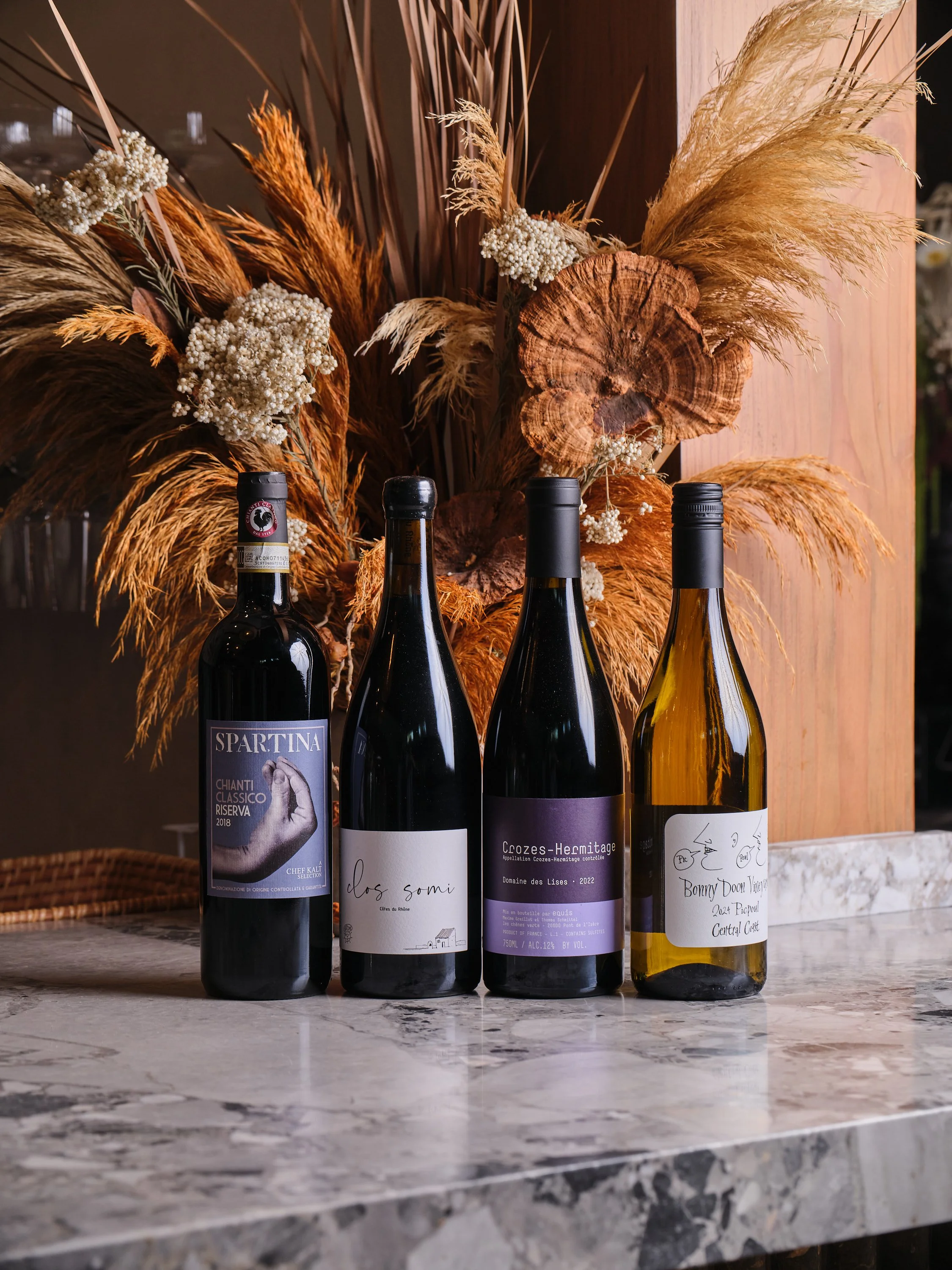 Four bottles of wine on a marble countertop with a decorative dried floral arrangement in the background.