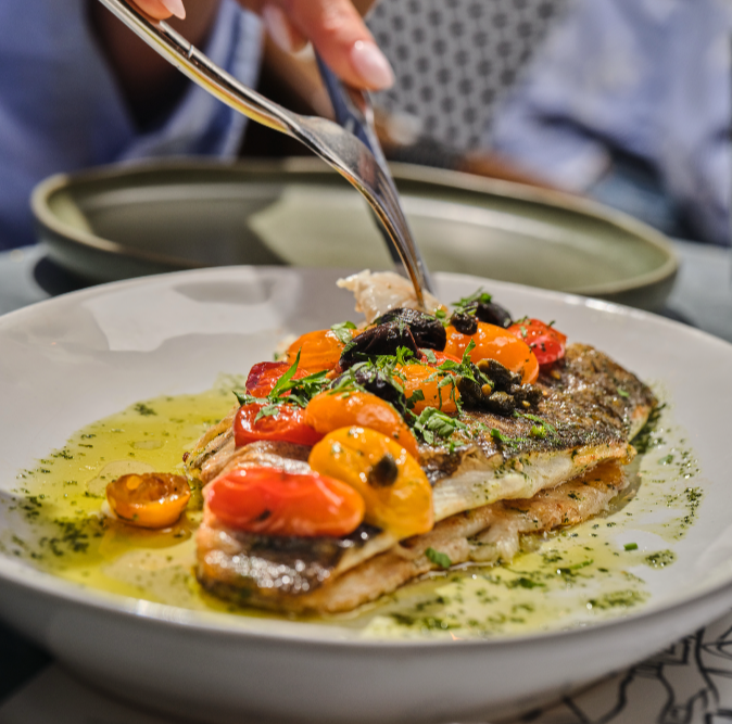 A plated dish of cooked fish topped with cherry tomatoes, herbs, and black olives, with a green sauce underneath.