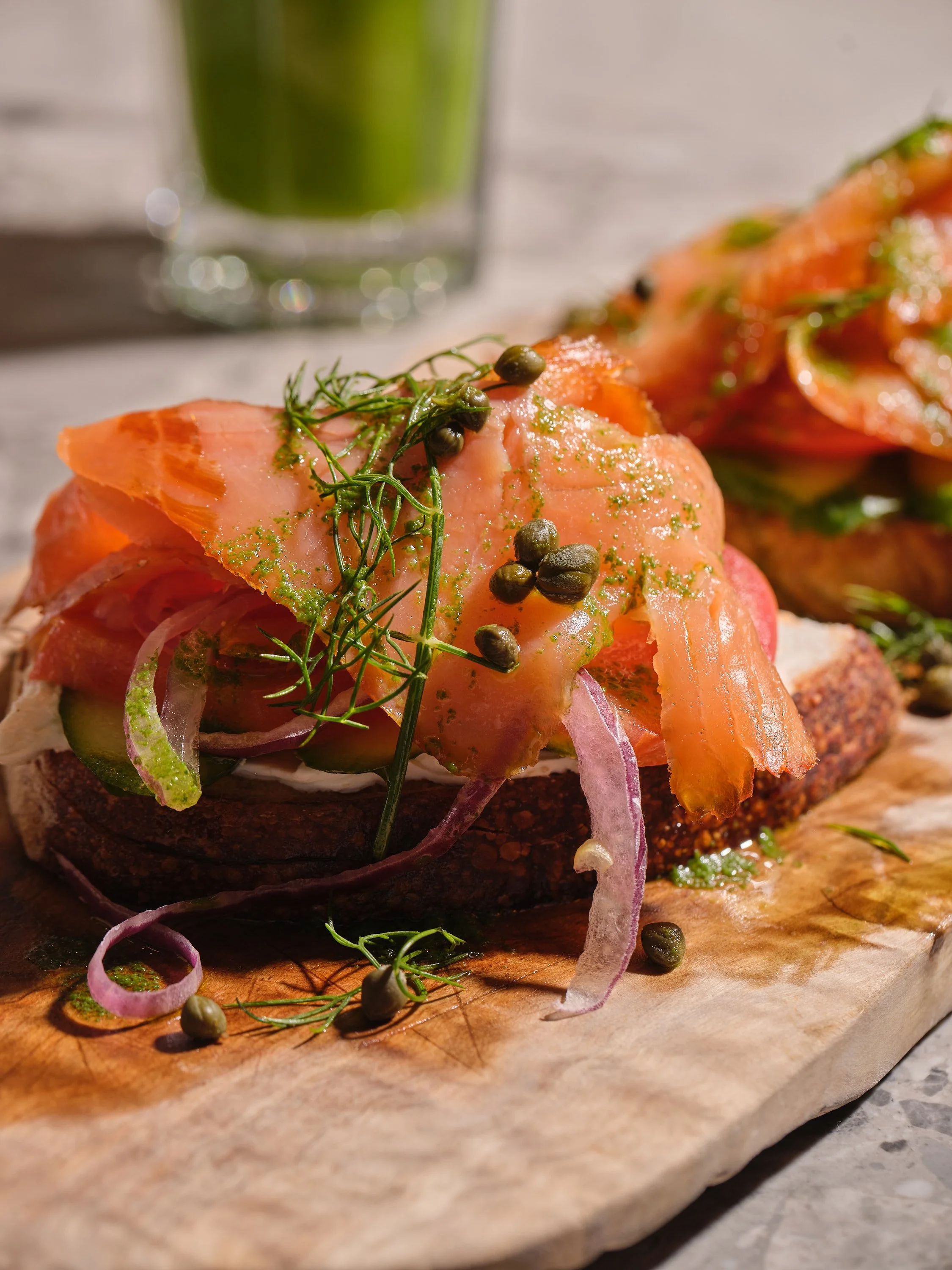 Close-up of smoked salmon on a toasted bagel with onion slices, cucumber, dill, capers, and green sauce on a wooden board with a glass of green juice in the background.