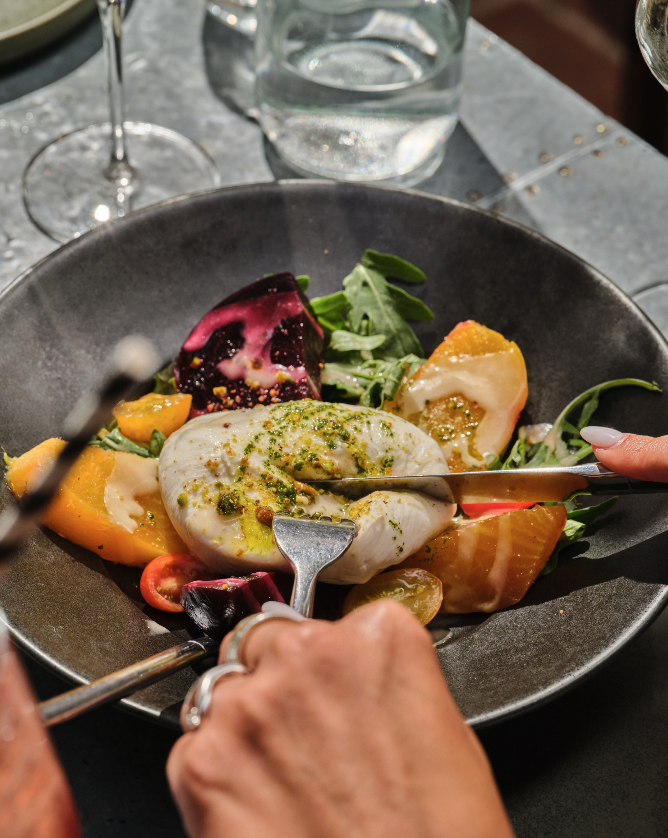A black plate with a fresh salad including yellow and red cherry tomatoes, beet, and arugula, topped with burrata cheese being drizzled with green pesto.