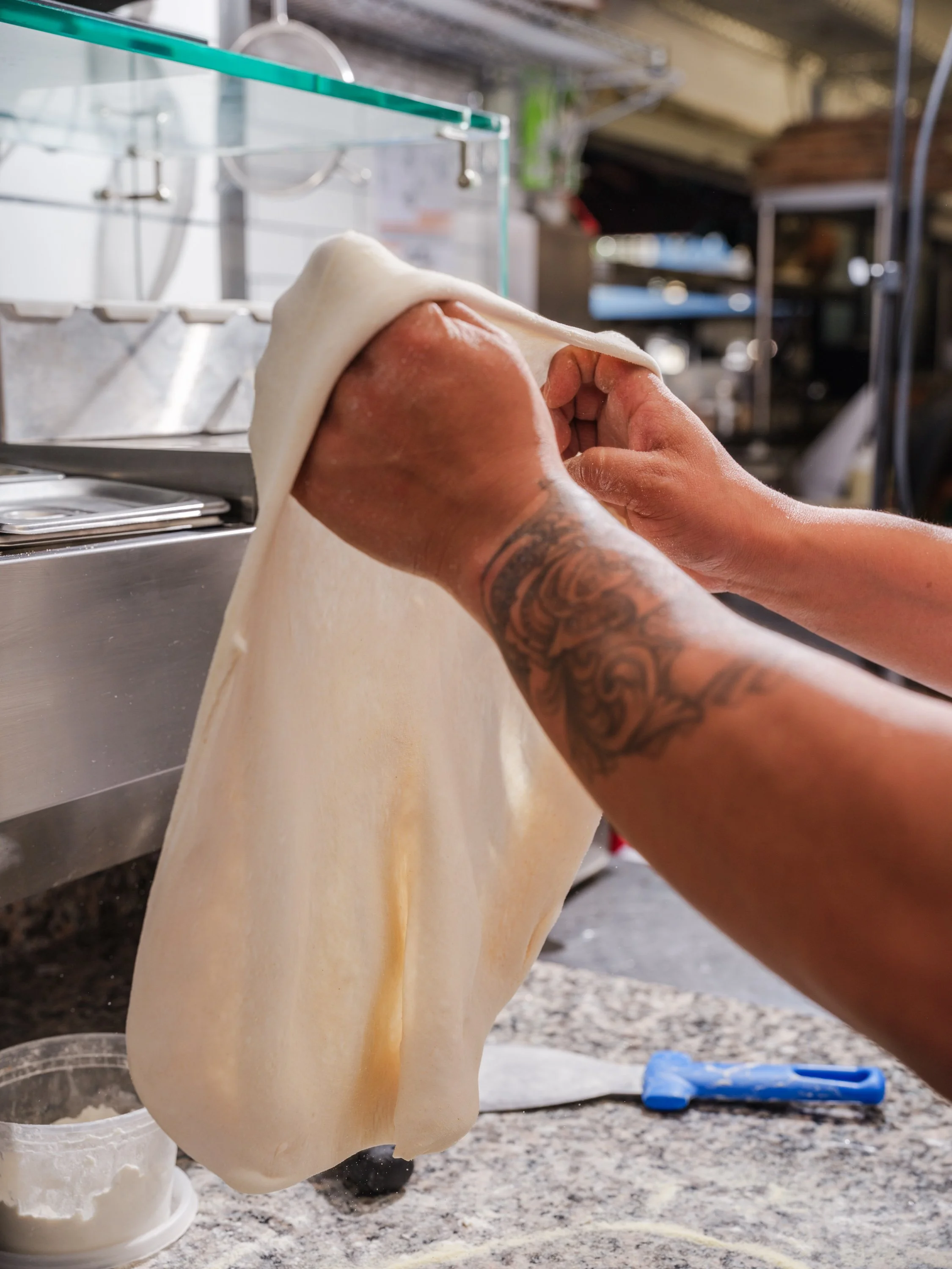 A person stretching pizza dough in a kitchen with stainless steel counters and shelves.