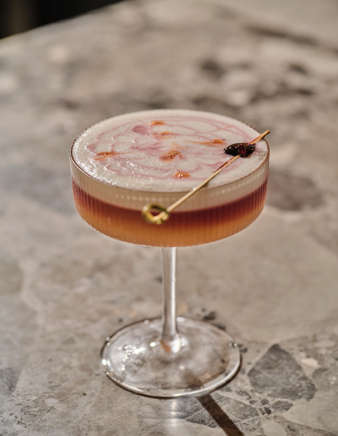Pink cocktail in a coupe glass garnished with a small skewer and a cherry.