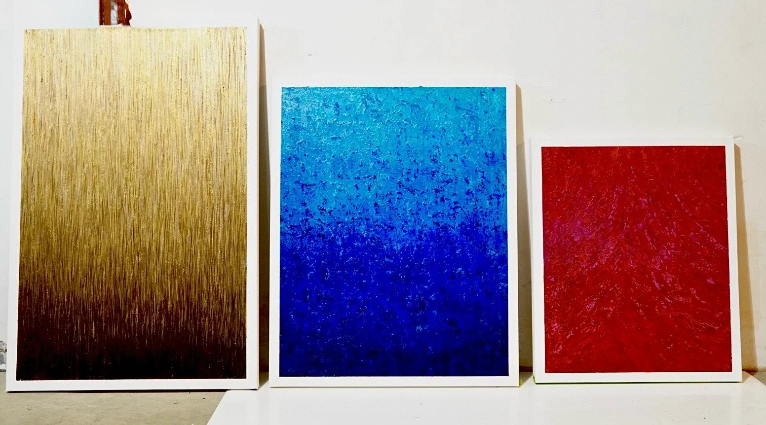 Three framed abstract paintings with gradient textures, a large gold piece on the left, a blue painting in the center, and a red painting on the right, against a white wall.