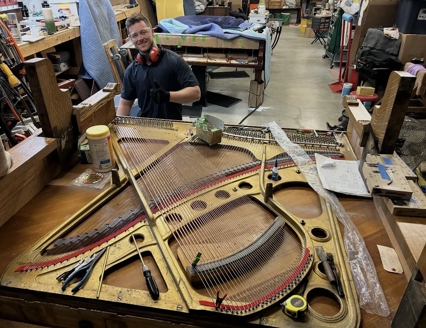 A young man wearing safety glasses, black gloves, and a black shirt giving a thumbs up at a workbench with an open piano, showing its strings and inner components, in a workshop environment.